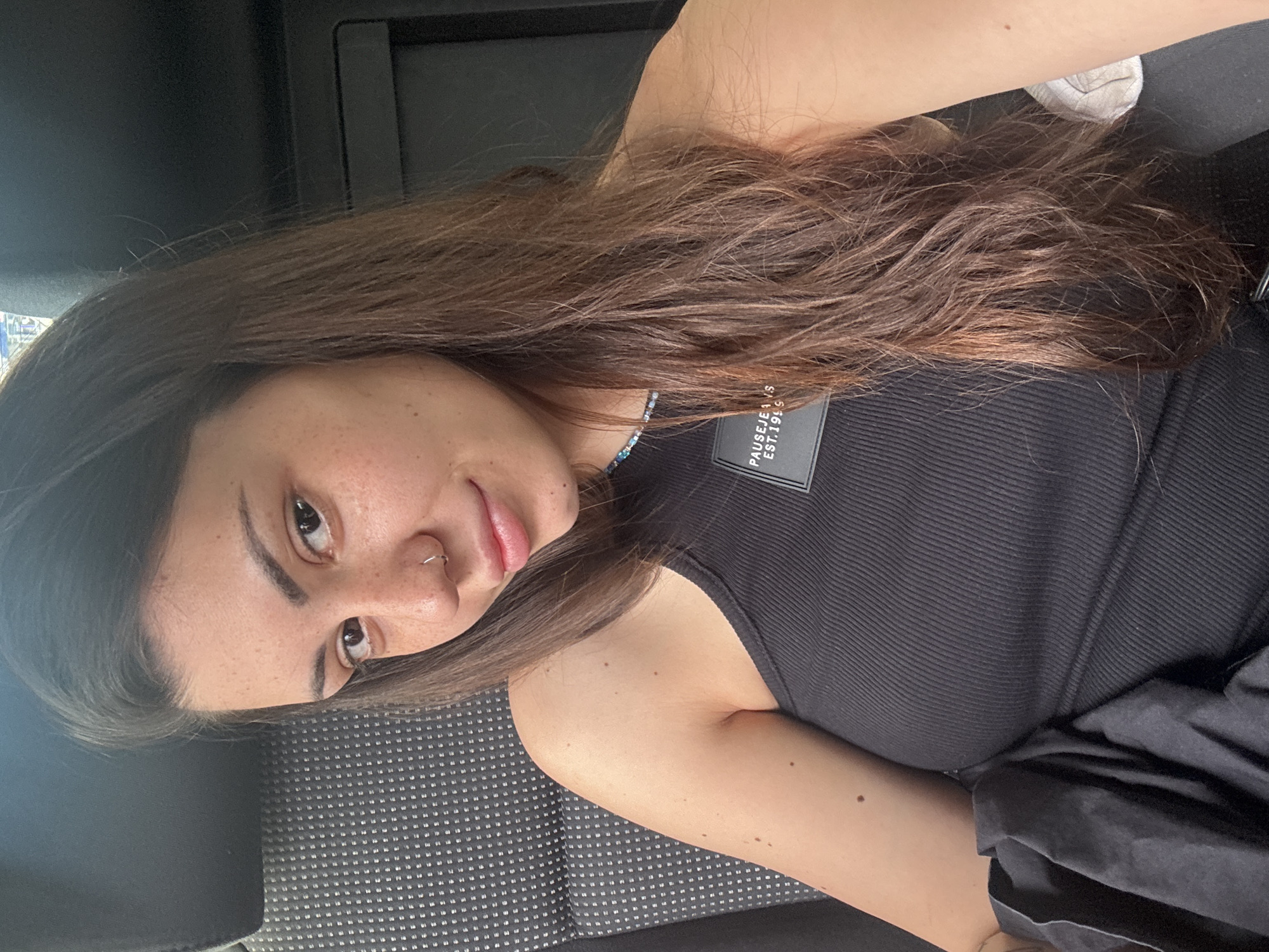A young woman is captured in a medium shot, rotated to appear upright, within a car interior, likely in Sofia, Bulgaria. She has long, flowing dark brown hair that partially covers her shoulders and chest. Her gaze is directed towards the viewer with a soft, neutral expression, possibly a faint smile. Her face features noticeable freckles across her nose and cheeks, and she has a silver ring piercing on her left nostril.
She is wearing a black ribbed tank top or camisole. A visible label on her top clearly displays the text "PAUSE JEANS" and "EST. 1999". Around her neck, she wears a delicate beaded necklace with varying shades of blue.
The background consists of the car's interior, identifiable by the patterned gray and black fabric of the seat she is resting against and the dark surfaces of the vehicle's cabin. The lighting suggests it is daytime, providing natural illumination to her face. She appears to be posing for a photograph or selfie in a casual setting. Her complexion is fair, and her arm is visible resting casually.