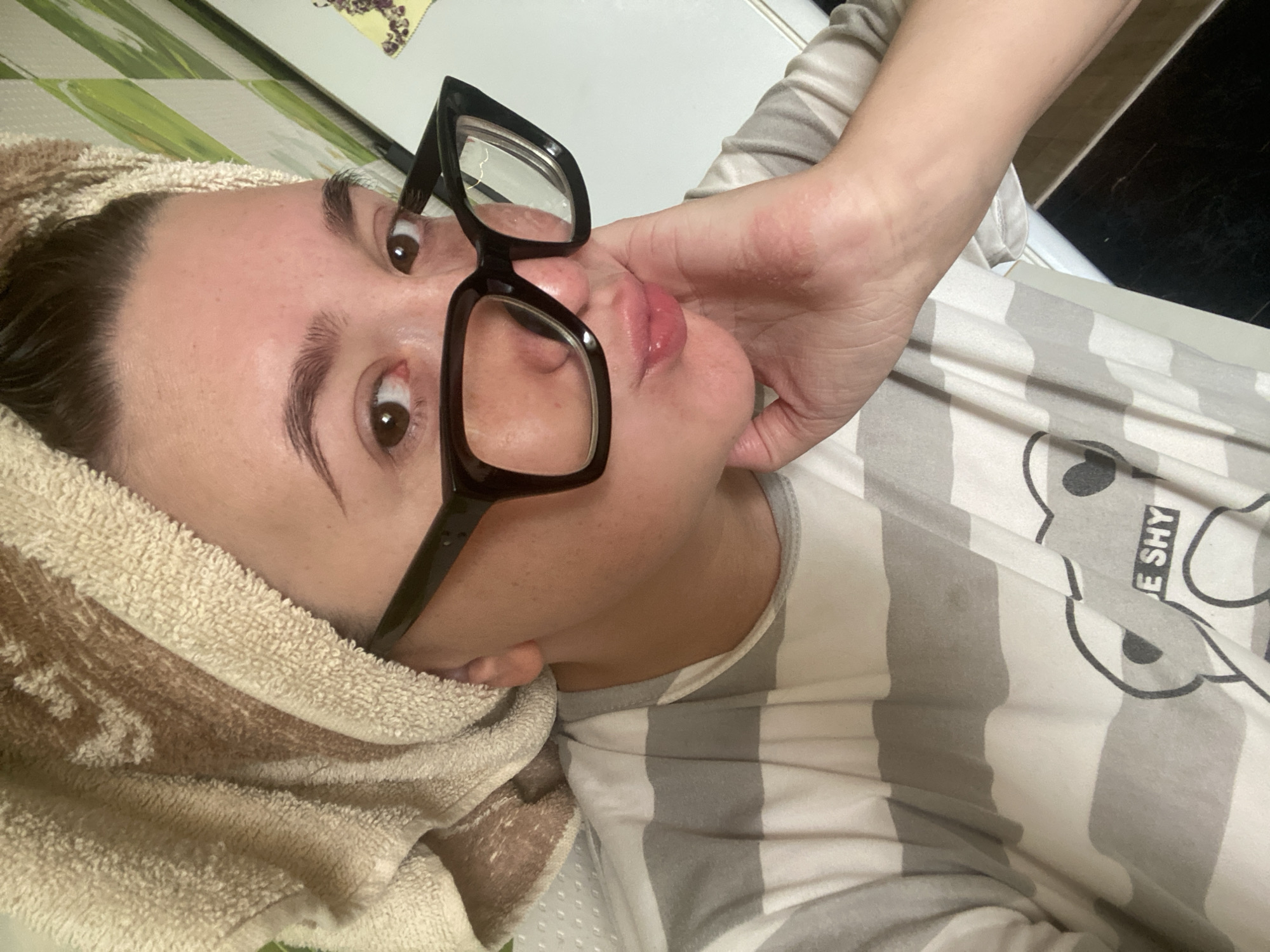 An adult individual, appearing to be female, is depicted from the chest up, positioned facing the viewer. The person has dark hair pulled back and wrapped in a light brown terry cloth towel. Large, rectangular-framed black eyeglasses are worn, with clear lenses. The eyes are brown and directed towards the camera. The lips are puckered in a kissing gesture, and the chin/jaw is resting on the back of the right hand. The skin on the face shows some minor redness, and there is visible redness or irritation on the back of the right hand and wrist. The individual is wearing a t-shirt with horizontal stripes of alternating white and light gray. A partial graphic design, including dark outlines of what appears to be a cartoon bear and the text "E SHY," is visible on the lower right portion of the shirt. The background features a textured white surface on the left, and a wall with vertical light green and white/beige stripes on the upper right. Below the striped wall is a white surface, possibly a refrigerator or cabinet, with a small decorative item featuring purple elements. A dark, possibly tiled, surface is visible in the lower right corner, separated by a thin white strip. The overall scene suggests an indoor domestic setting.
