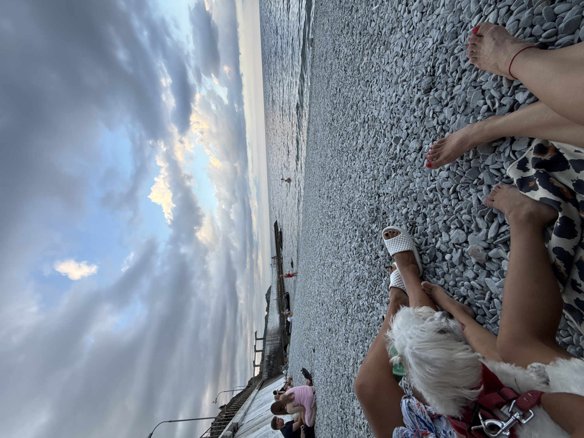 In Partenit, Ukraine, a pebble beach stretches towards the Black Sea under a dramatic cloudy sky. In the foreground, two individuals lounge on the pebbles, with their legs visible. One person's legs are adorned with white slides and a small white dog rests on their lap. The other person's legs are also visible, with one foot resting on the other and a red anklet. The background shows a concrete embankment with steps leading down to the sea, and several people are scattered across the beach and in the water. The overall scene depicts a relaxed day at the seaside.
