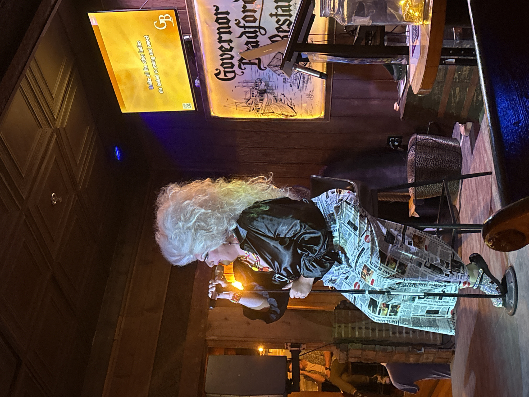 In Provincetown, Massachusetts, a drag performer with voluminous white hair, wearing a black satin blouse and pants printed with newspaper clippings, sits on a stool and holds a microphone to their mouth, appearing to sing. A large television screen behind the performer displays lyrics: "Unlike all the other ladies she looked so young and sweet," along with a logo. To the right of the television, a lit sign reads "Governor Bradford Restaurant." The room features wooden walls and a dark, textured ceiling. The scene suggests an indoor performance venue.