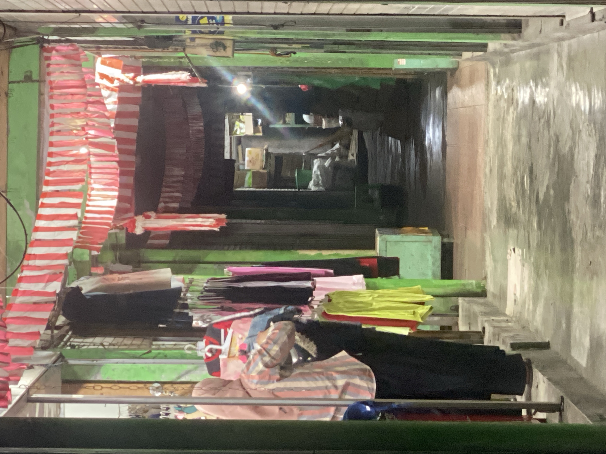The image depicts a narrow, dimly lit alleyway in Ambarketawang, Indonesia, likely a market or commercial area. The walls of the alley are painted a vibrant green. Red and white striped fabric banners are hung overhead, creating a festive or decorative atmosphere. On the left side of the alley, a person wearing a pink hijab and a striped shirt is standing, appearing to browse or handle clothing that is hanging on a rack. Several garments, including dresses and tops in various colors such as pink, black, yellow, and red, are displayed on hangers. The ground of the alley appears to be wet and reflects the light. Further down the alley, the scene opens up into a darker area with storage shelves and what appear to be boxes and bags. A single light source illuminates the deeper part of the alley, casting a glare.