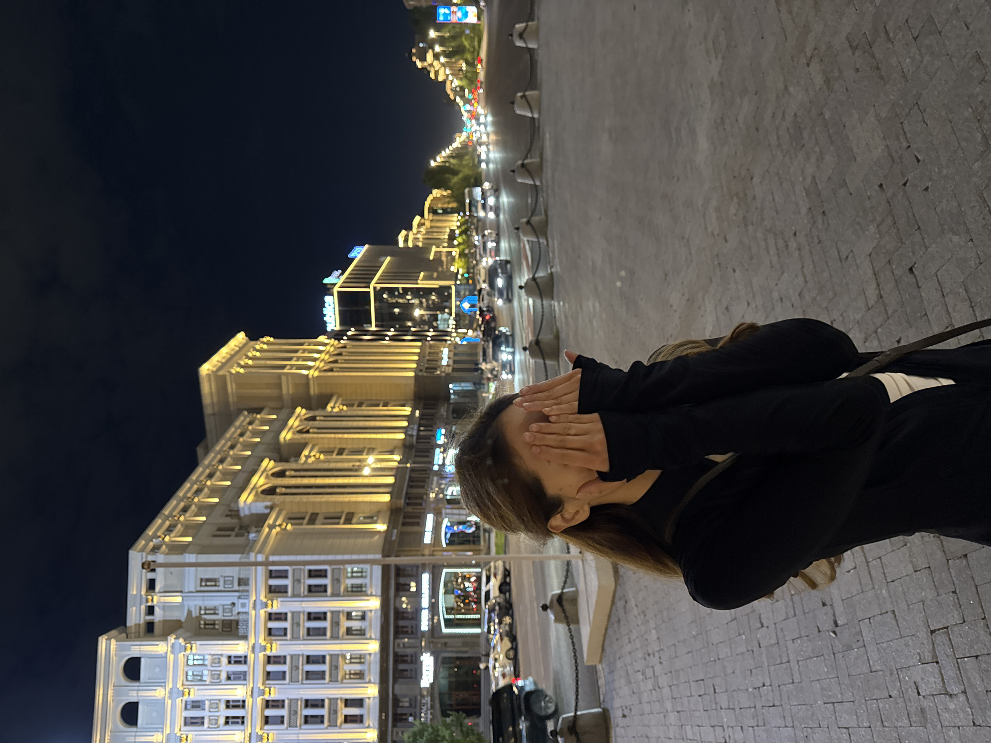 A medium shot captures a young woman standing in a city square at night, covering her face with her hands. She is wearing a black top with long sleeves and a white undershirt. Her hair is light brown and falls past her shoulders. She is standing on a paved surface, and a cobblestone walkway is visible to her left.
In the background, a wide street is lined with modern buildings illuminated by warm yellow lights. The architecture suggests a European style, with columns and arched windows. A prominent building in the mid-ground features a neon sign that appears to read "Republic." Cars are driving on the street, their headlights and taillights creating streaks of light. Streetlights line the road, casting a warm glow on the scene. The sky above is dark and clear. The overall impression is one of a well-lit urban environment at night.
The scene is identified as the Republic Square in Astana, Kazakhstan.