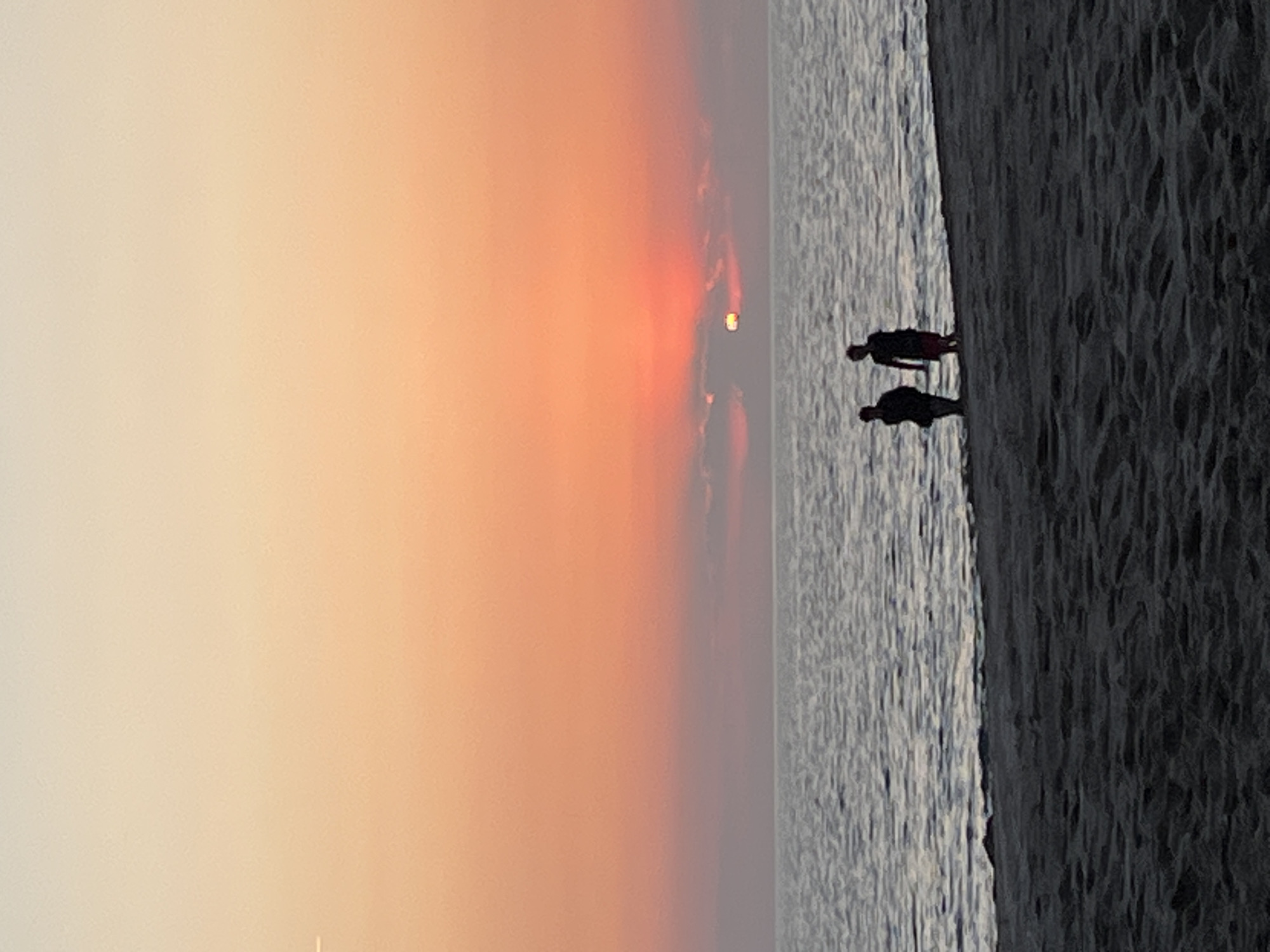 This photograph depicts a twilight or sunset scene at a beach in Alanya, Turkey. The image is oriented vertically. On the right side of the frame is the dark sand of the beach. Adjacent to the sand is the ocean, reflecting some of the light from the setting sun. Two silhouetted human figures are walking on the wet sand at the edge of the water. The left side of the image is dominated by the gradient-colored sky, fading from light at the top to a darker orange near the sun, which is setting on the horizon. The scene appears to be calm and peaceful.