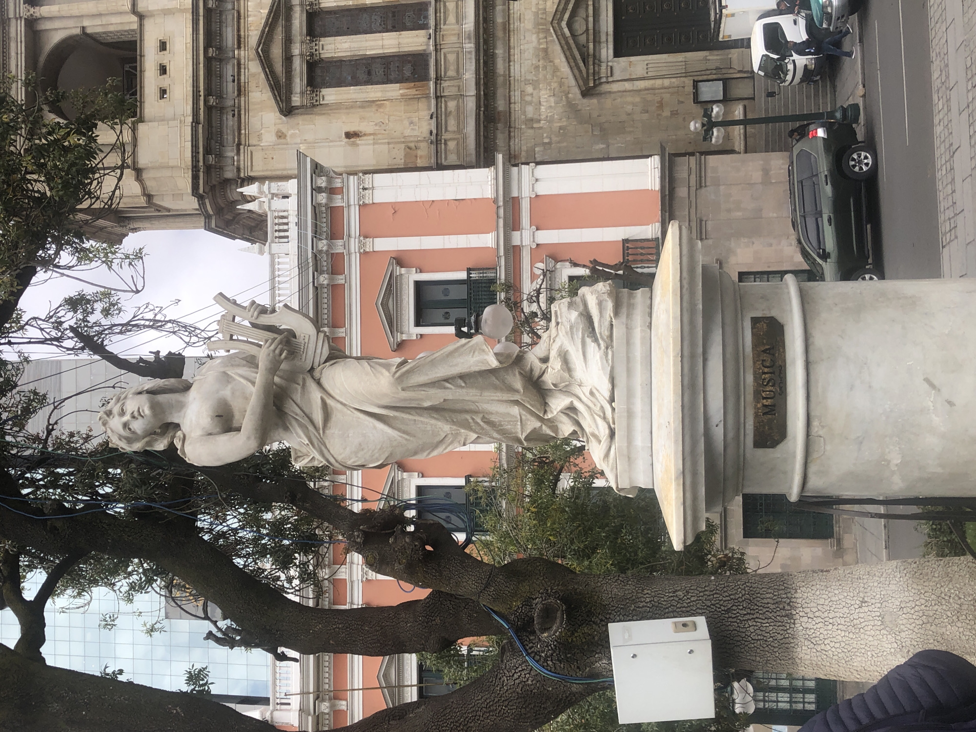 A marble statue of a woman playing a lyre stands on a pedestal in a public square, identified by a plaque reading "MÚSICA." The statue is located in La Paz, Bolivia, in front of an old building with a light pink and white façade. A large tree stands next to the statue, obscuring part of the view. Modern cars and people can be seen on the adjacent street.