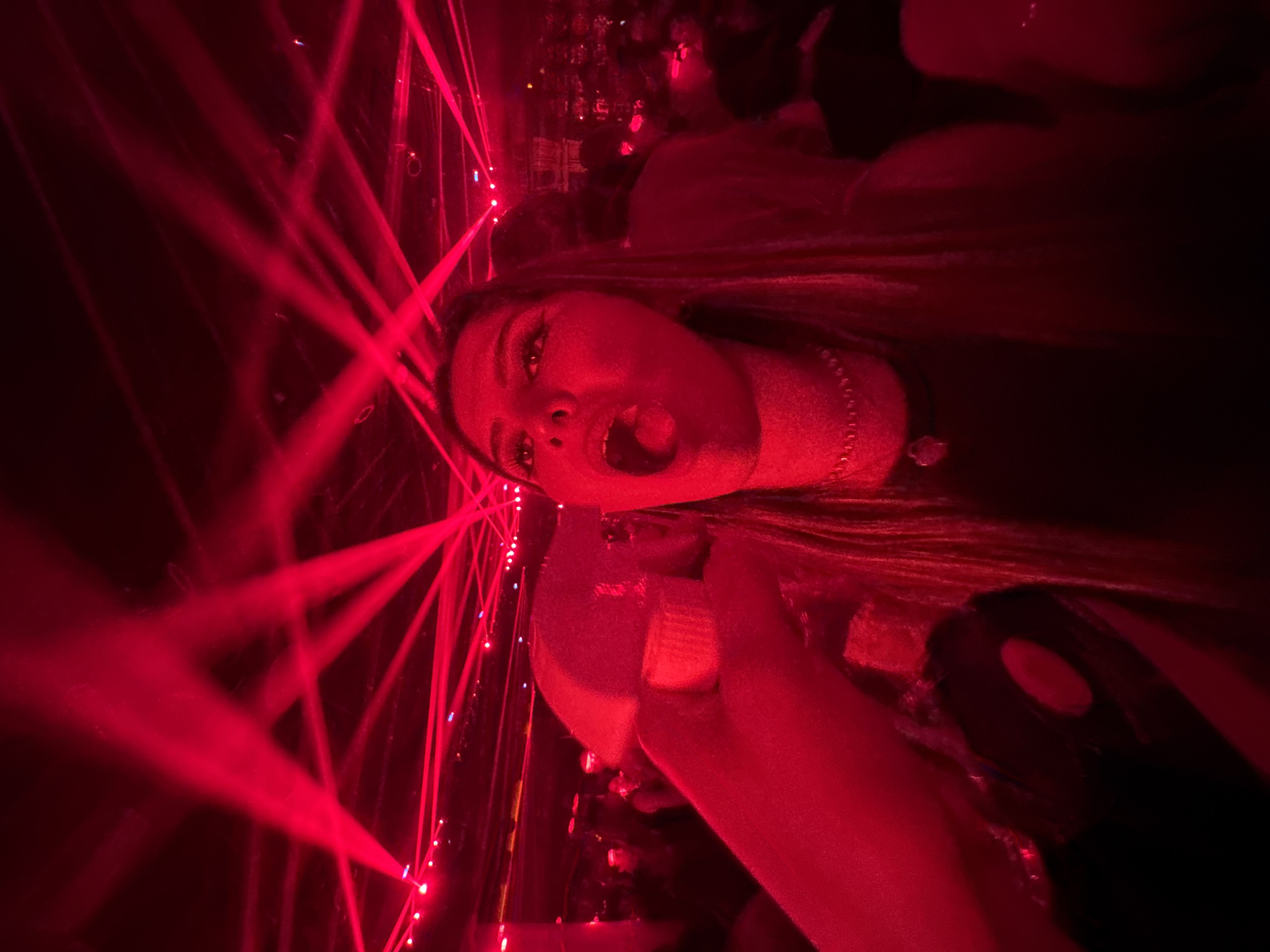 This media file captures a vibrant, high-energy scene featuring a woman in a dark, club-like environment, intensely illuminated by numerous red laser beams. The image is oriented with the woman's head tilted to the right, appearing to be on her side.
The primary subject is a woman with long, dark hair and a beaded necklace. Her face is brightly lit by the red lights, showing her eyes wide open and her mouth open as if shouting, singing, or reacting with excitement to the surrounding activity. Her expression conveys a sense of exhilaration and engagement with the event. In her hand, visible in the lower right, she holds what appears to be a drink in a clear container.
The setting is an indoor location, likely a nightclub, concert, or party, characterized by its low light levels and the prominent use of red laser lights that crisscross the frame, creating striking visual effects and an energetic atmosphere. Blurred shapes in the background suggest the presence of other people or stage elements, further emphasizing the event context. The time of day is clearly night. No discernible text is visible within the image. The location context provided is San Bernardino Tlaxcalancingo, Mexico.