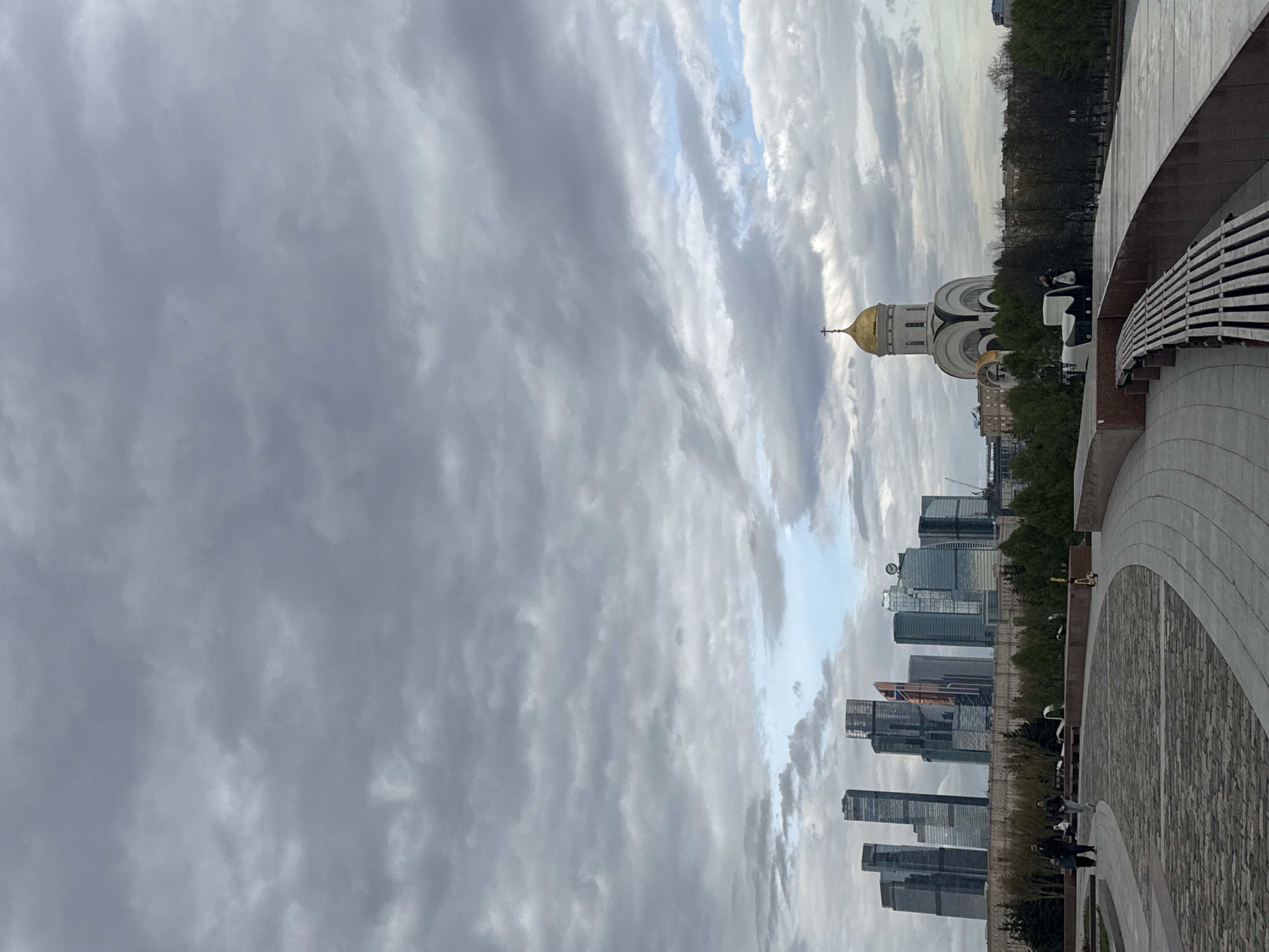 The image shows a view of Moscow, Russia. The foreground is a stone paved area with a curved concrete structure. The background shows a cityscape with modern high-rise buildings and the Cathedral of Christ the Savior with its golden dome. The sky is overcast with heavy clouds. There are people visible in the paved area and near the edge of the structure.
