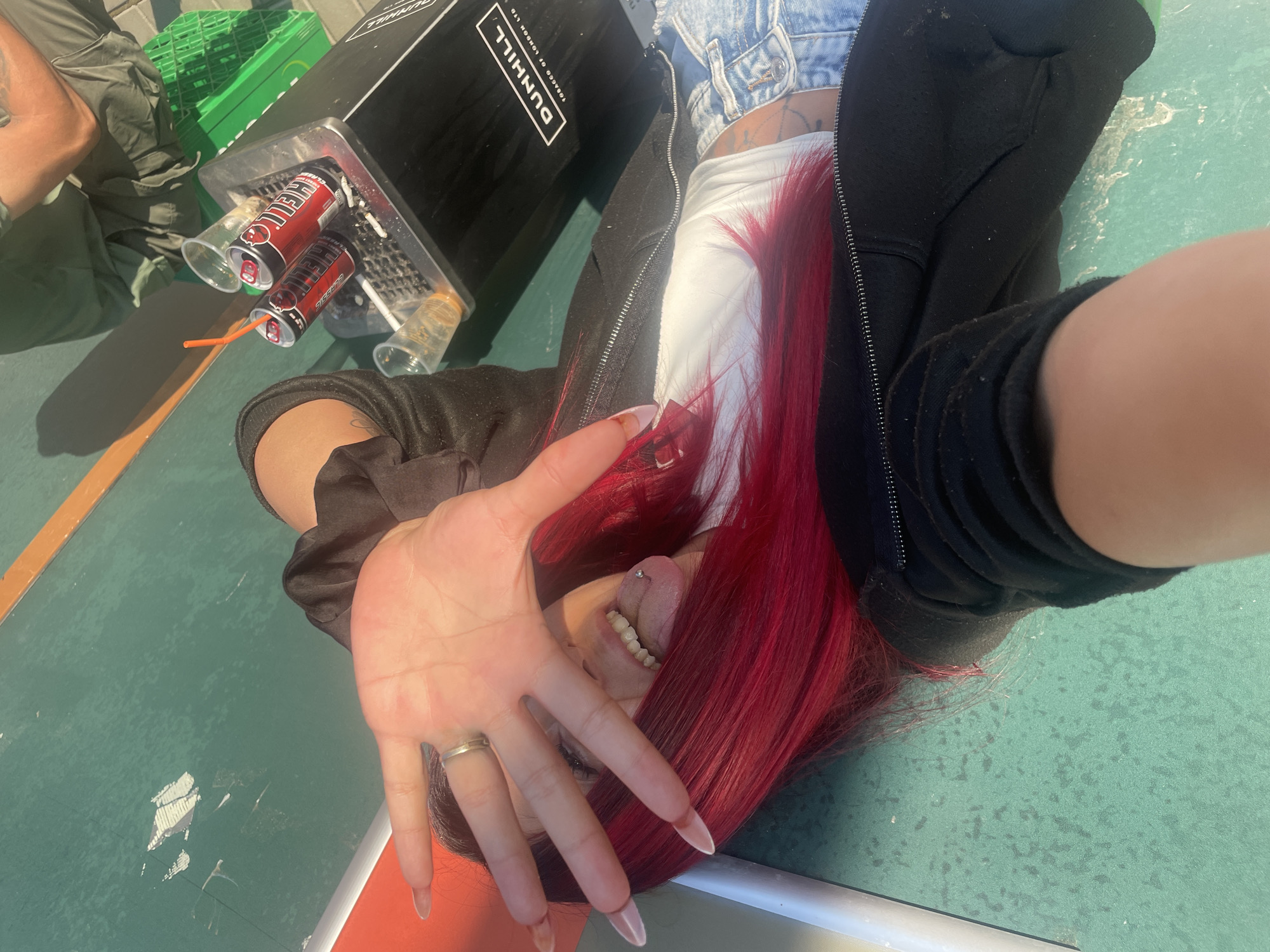 An overhead medium shot captures a young woman with vibrant red hair lying down on a green, textured surface. She is wearing a black zip-up hoodie over a white crop top and light blue distressed denim shorts. Her right hand is raised, partially obscuring her face, with her tongue sticking out and a silver ring on her left index finger. A silver tongue piercing is visible. To her right, on a black bin with "DUNHILL TOBACCO OF LONDON LTD" printed on it, are two red cans labeled "HELL ENERGY" and two clear plastic cups, one with an orange straw. Cigarette butts are present in the bin's ashtray. The green surface appears to be an outdoor space, possibly a court or paved area, with a hint of an orange line and a white structure in the background. The lighting suggests it is daytime. This scene appears to be captured in a public area within the city of Sofia, Bulgaria.