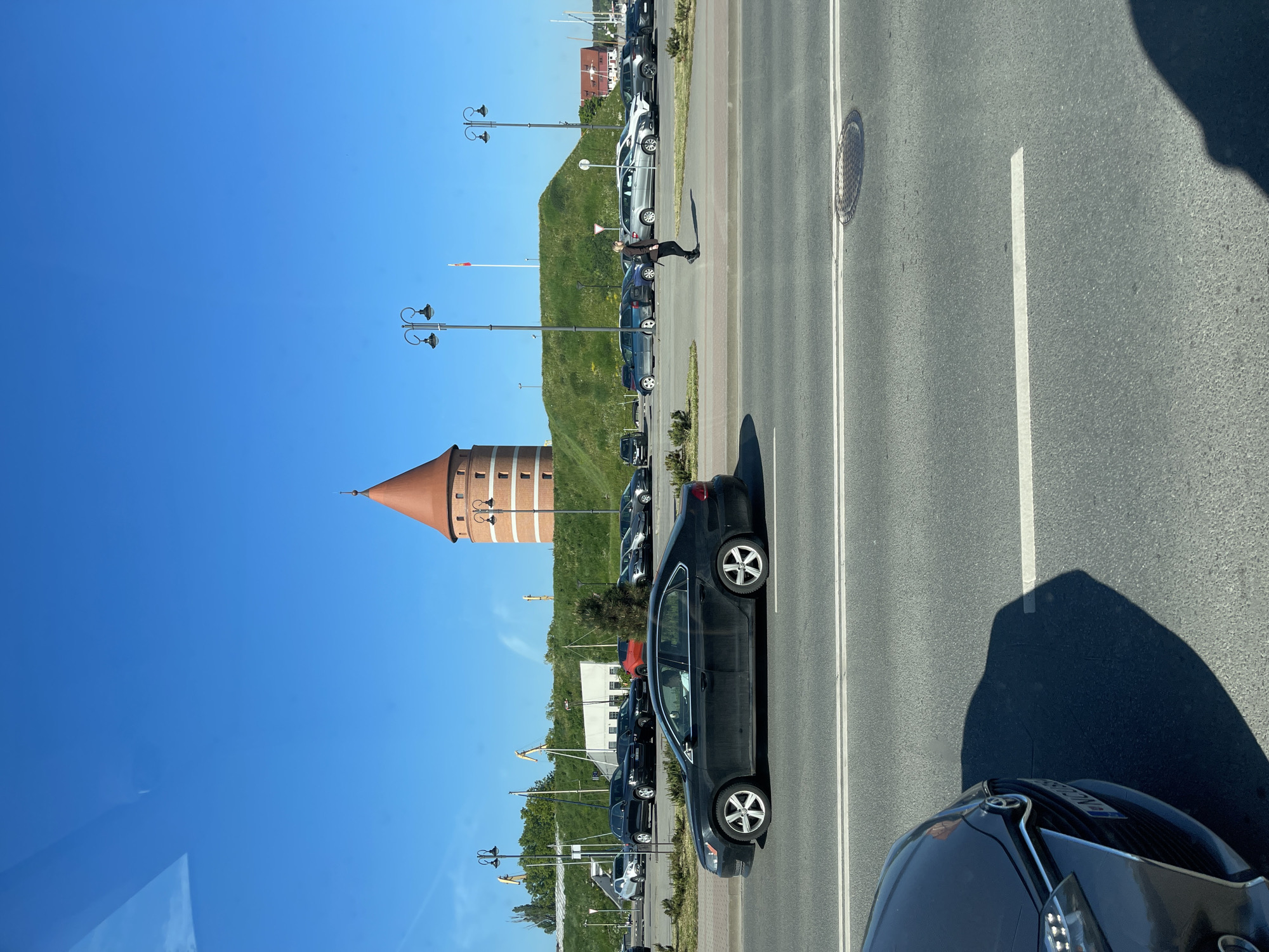 The image depicts a street scene with a view of a tower. In the foreground, there is a road with cars parked along the side. A dark-colored car is prominently positioned on the right side of the road, and parts of another car are visible in the bottom right corner.
Across the road, a row of parked cars lines the street, with a grassy hill and lampposts behind them. In the distance, a prominent brick tower with a conical roof rises against a clear blue sky. A person is walking on the sidewalk. The location is Unknown, Unknown.