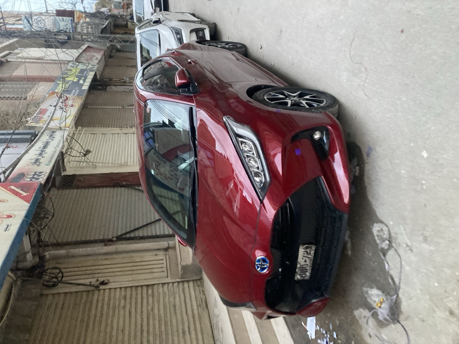 A red Toyota Aqua hybrid car is parked along a sidewalk, with a white car ahead. The red car's front is visible, including the headlights, grill, and license plate with "800-729" on it. Buildings and shops are visible on the left side, with metal doors and signboards. The image appears to be taken at an angle. There is some debris on the sidewalk.