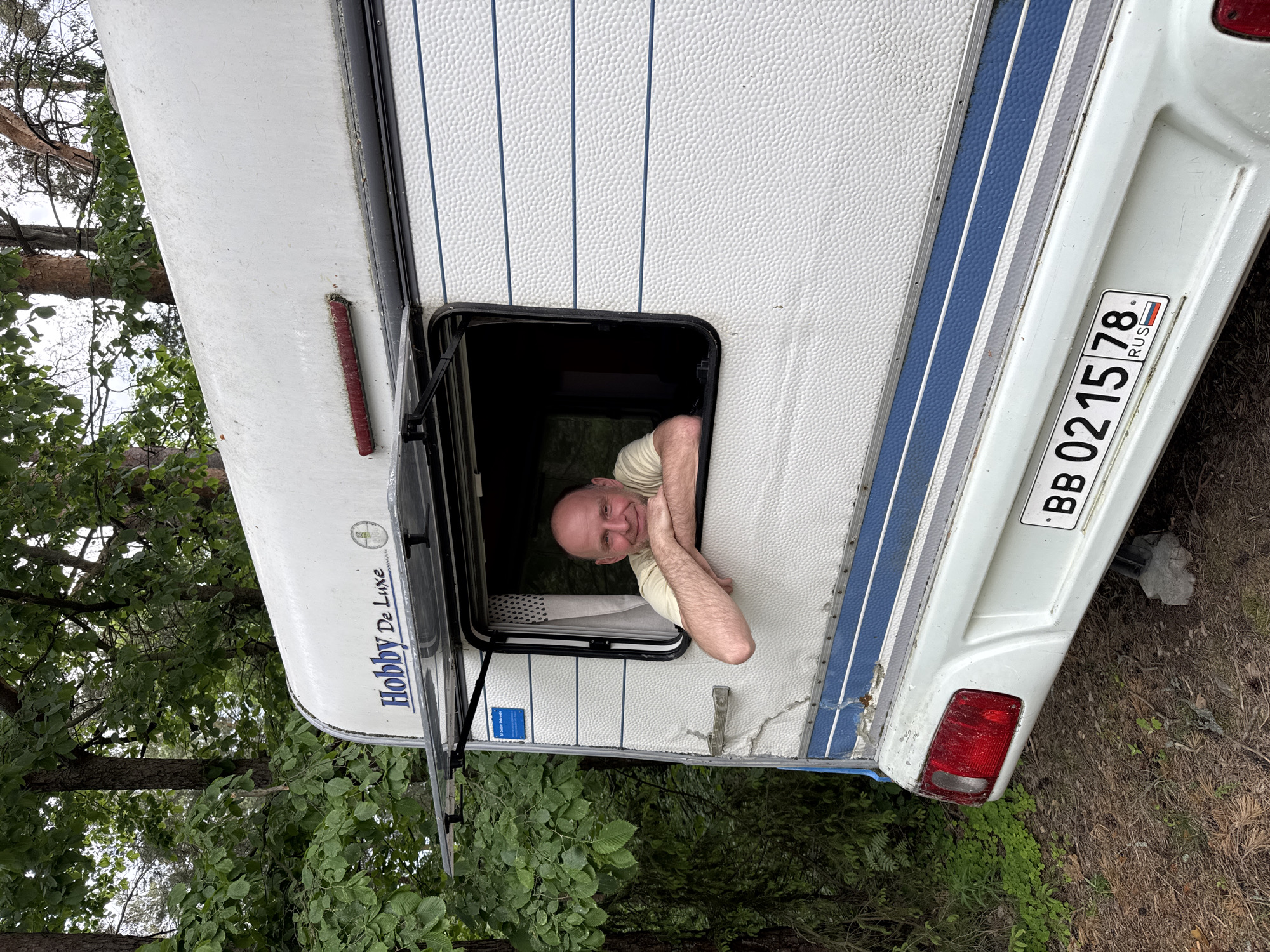 An adult male is prominently featured, leaning out of the open window of a white Hobby De Luxe camper van, which is stationary in a wooded outdoor setting in Fort-Krasnaya Gorka, Russia. The man, with a bald head and light stubble, wears a pale yellow short-sleeved t-shirt and smiles directly at the viewer, resting his forearms on the window's lower frame.
The camper van displays a textured white exterior with two blue horizontal stripes along its lower half and "Hobby De Luxe" branding on its upper rear. A dark red horizontal light bar is visible near the roofline, and a rectangular blue sticker is affixed to the side above the window. The vehicle exhibits signs of wear, including dirt accumulation, minor surface damage, and a weathered taillight on the lower left. A Russian license plate, "BB 0215 78 RUS," is mounted centrally on the rear bumper, with "78" indicating the Saint Petersburg region. A grey support block is positioned beneath the rear bumper.
The surrounding environment consists of dense green deciduous trees and foliage in the background, suggesting a natural, possibly forested area. The ground beneath the camper is composed of dirt and scattered organic debris, with sparse green ground cover. The lighting suggests daytime conditions, with diffused illumination.