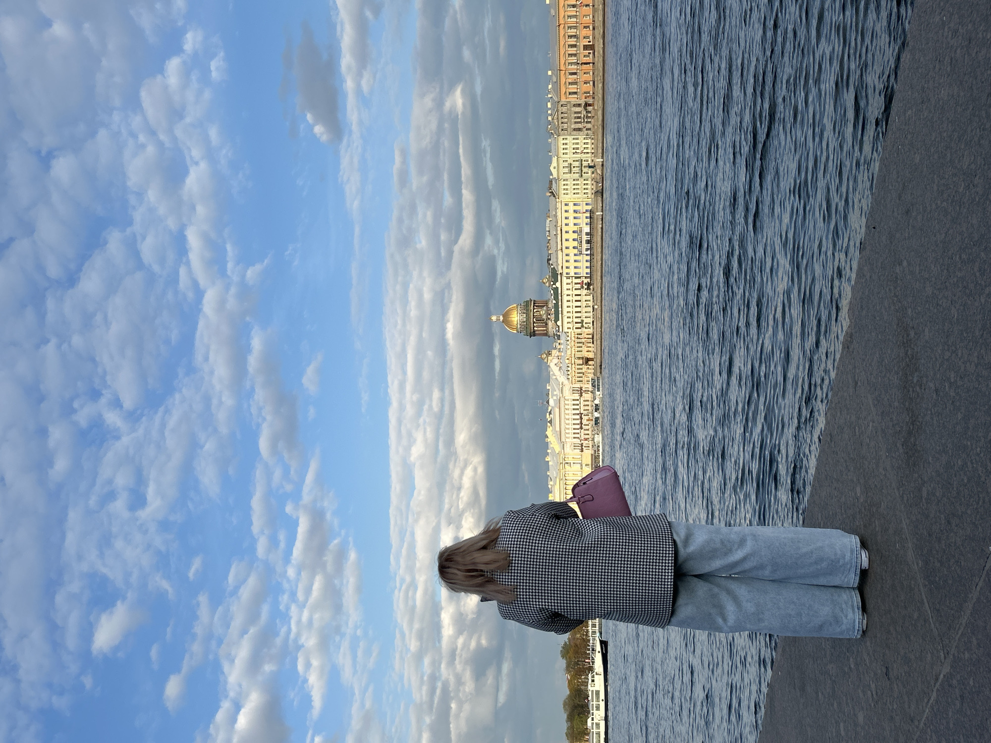 The photograph is oriented sideways and depicts a woman standing on a concrete embankment facing the Neva River and the cityscape of Saint Petersburg, Russia. The woman is seen from behind. She has long blonde hair and is wearing light blue jeans and a black and white houndstooth-patterned coat. She carries a purple purse. Across the river, the skyline is visible with buildings that feature traditional Russian architecture, including Saint Isaac's Cathedral, easily identified by its large golden dome. The sky is partly cloudy with a mix of light blue and white clouds. The water of the Neva River is choppy. The scene appears to be taken during daylight.