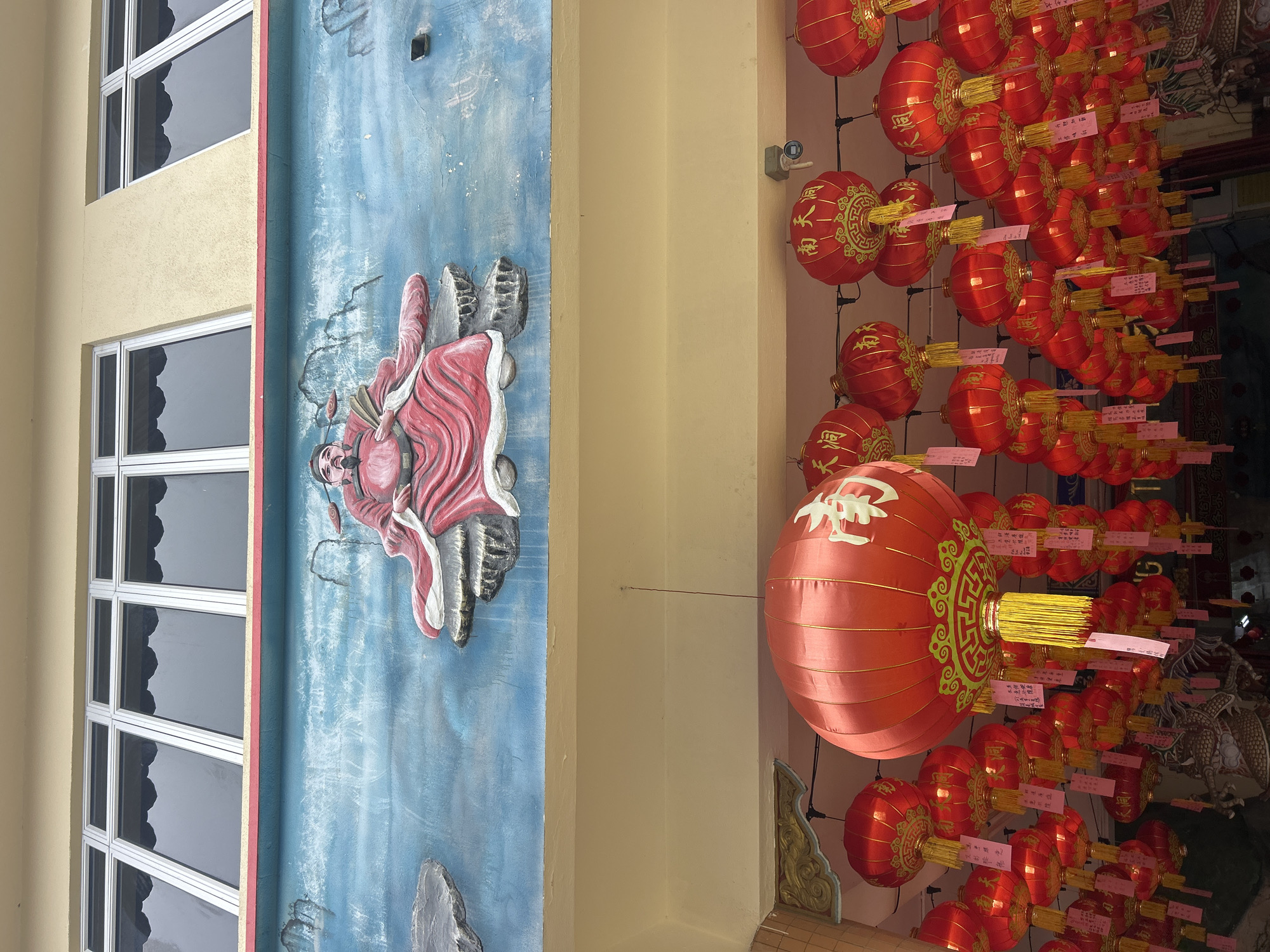 The image captures an exterior view of a building facade adorned with a blue mural and red lanterns. Above the mural, a row of windows with aluminum frames is visible. The mural depicts a seated figure in traditional red attire, holding a fan, against a blue backdrop suggestive of clouds or water. Below the mural, numerous red, cylindrical lanterns are strung, many bearing Chinese characters in gold. Some lanterns have yellow tassels and pink tags attached. The scene appears to be a cultural or religious site, possibly a temple, and is located in Ipoh, Malaysia, as indicated by the signage on the lanterns. The overall atmosphere is festive and cultural, with a focus on traditional Chinese elements.