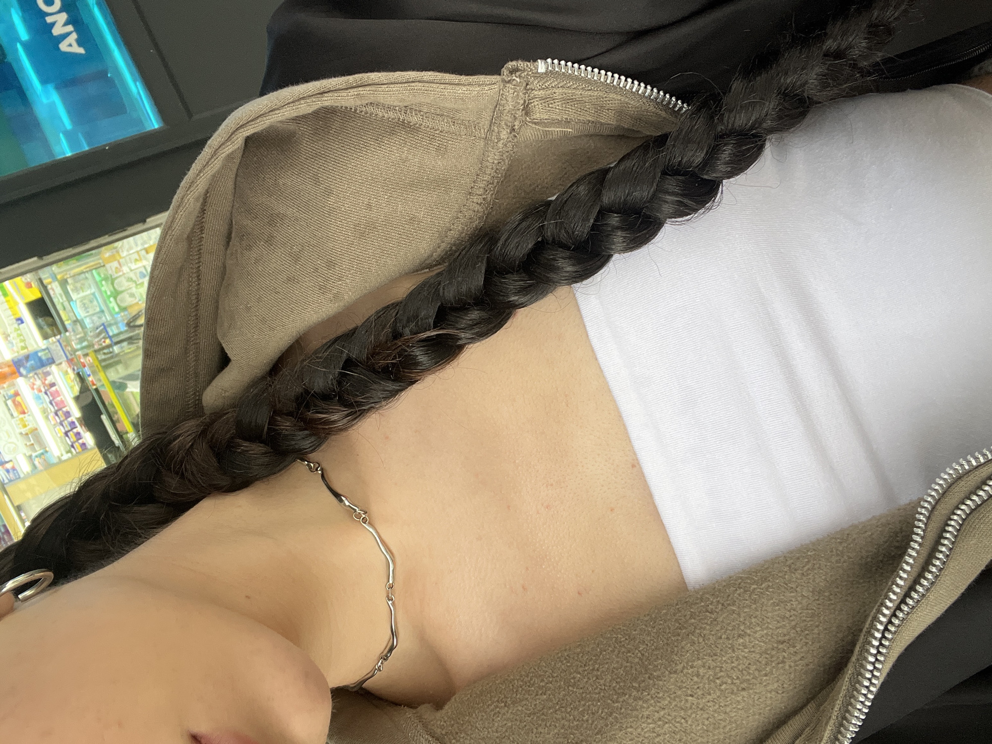 The image captures a close-up, downward-angled view of a person's upper torso, neck, and a portion of their head. The individual has long, dark hair styled into a thick braid that drapes across their chest. They are wearing a plain white tube top or tank top, partially covered by an unzipped, olive-green or khaki-colored hoodie or jacket made of a textured, possibly fleecy material. A silver, irregular-linked chain necklace adorns their neck, and a silver ring is visible on what appears to be their left hand, near the bottom left corner.
The background suggests an indoor retail environment, likely a convenience store or similar shop, in Ternopil, Ukraine. Behind the person, shelves stocked with various products are visible, illuminated by overhead lights. A bright blue, rectangular sign with the partial text "ANC" is prominently displayed in the upper left background. The overall lighting is artificial. There is no specific activity depicted other than the person being present in this setting. Small red spots are visible on the skin of the neck/chest area. The image focuses on textures and details of clothing and accessories against the retail backdrop.