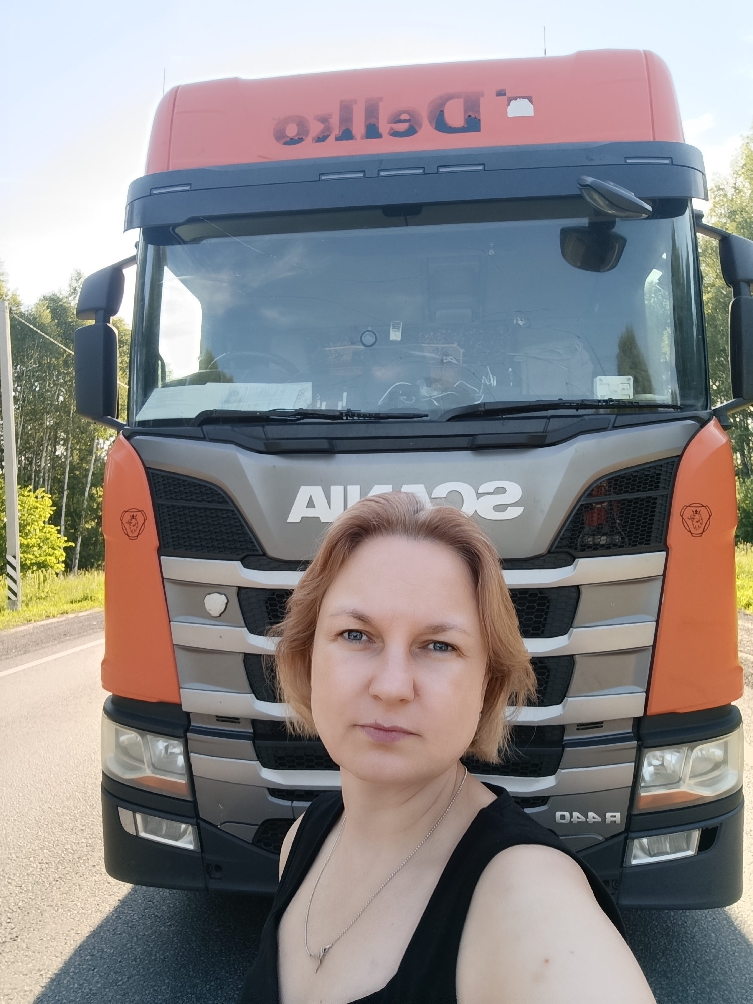 This media file captures a selfie of a woman with blonde hair in front of a large orange and grey semi-truck. The truck's grille prominently displays the "SCANIA" logo in large silver lettering. The truck is parked on the side of a paved road, with a grassy verge and trees visible in the background. The sky is bright and clear, suggesting a sunny day, likely in the late morning or afternoon. The woman is wearing a black tank top and a silver necklace. Her expression is neutral. The truck has "DeIko" written in blue lettering on its orange upper panel, although some of the letters appear to be obscured or damaged. The location is identified as L'vovo, Russia. The scene appears to be a roadside stop during what seems to be a journey or a work-related pause for the truck driver.