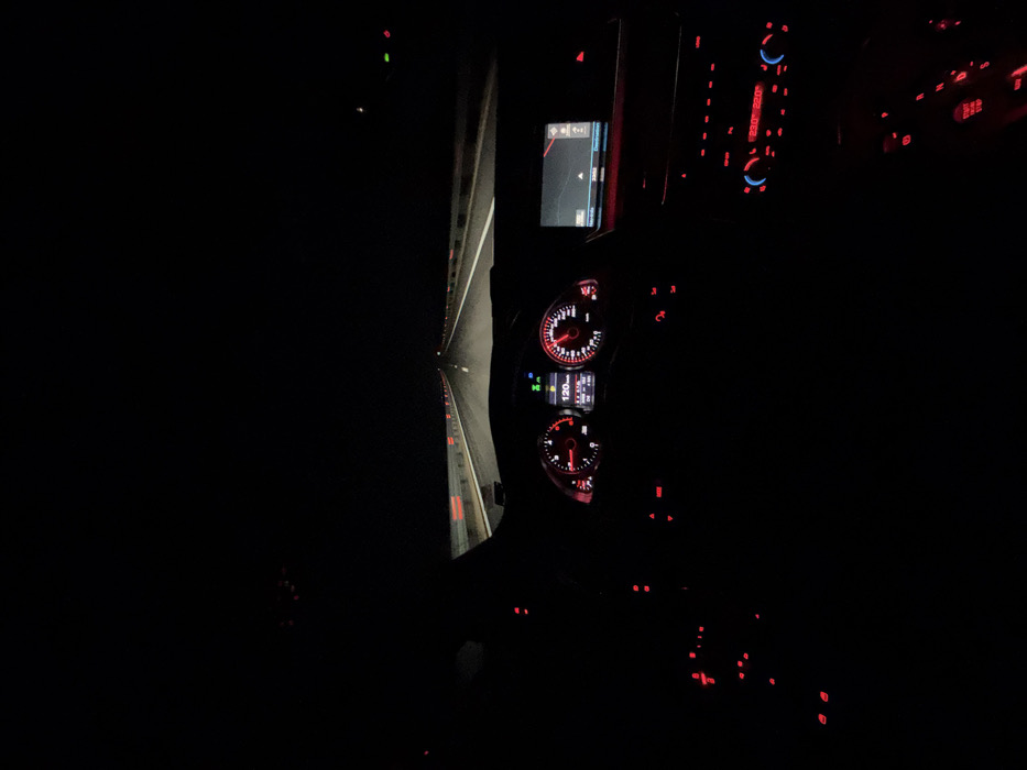 A nighttime scene is depicted from the driver's perspective inside a vehicle traveling on a multi-lane highway near Gradishte, Bulgaria. The road ahead is illuminated by the vehicle's headlights, revealing a straight stretch of asphalt with white dashed lane markings. Red-reflective markers are visible on the guardrails lining both sides of the road. The sky above is uniformly dark.
Inside the vehicle, the illuminated dashboard features an instrument cluster with a speedometer indicating 120 km/h and a tachometer showing approximately 2000 RPM. A central digital display within the cluster presents additional vehicle data. To the right, an infotainment screen displays a navigation map. Below this, a climate control panel shows temperature settings of 23.0°C and 20.0°C, surrounded by numerous red-illuminated buttons on the center console and dashboard. No individuals are visible within the frame.