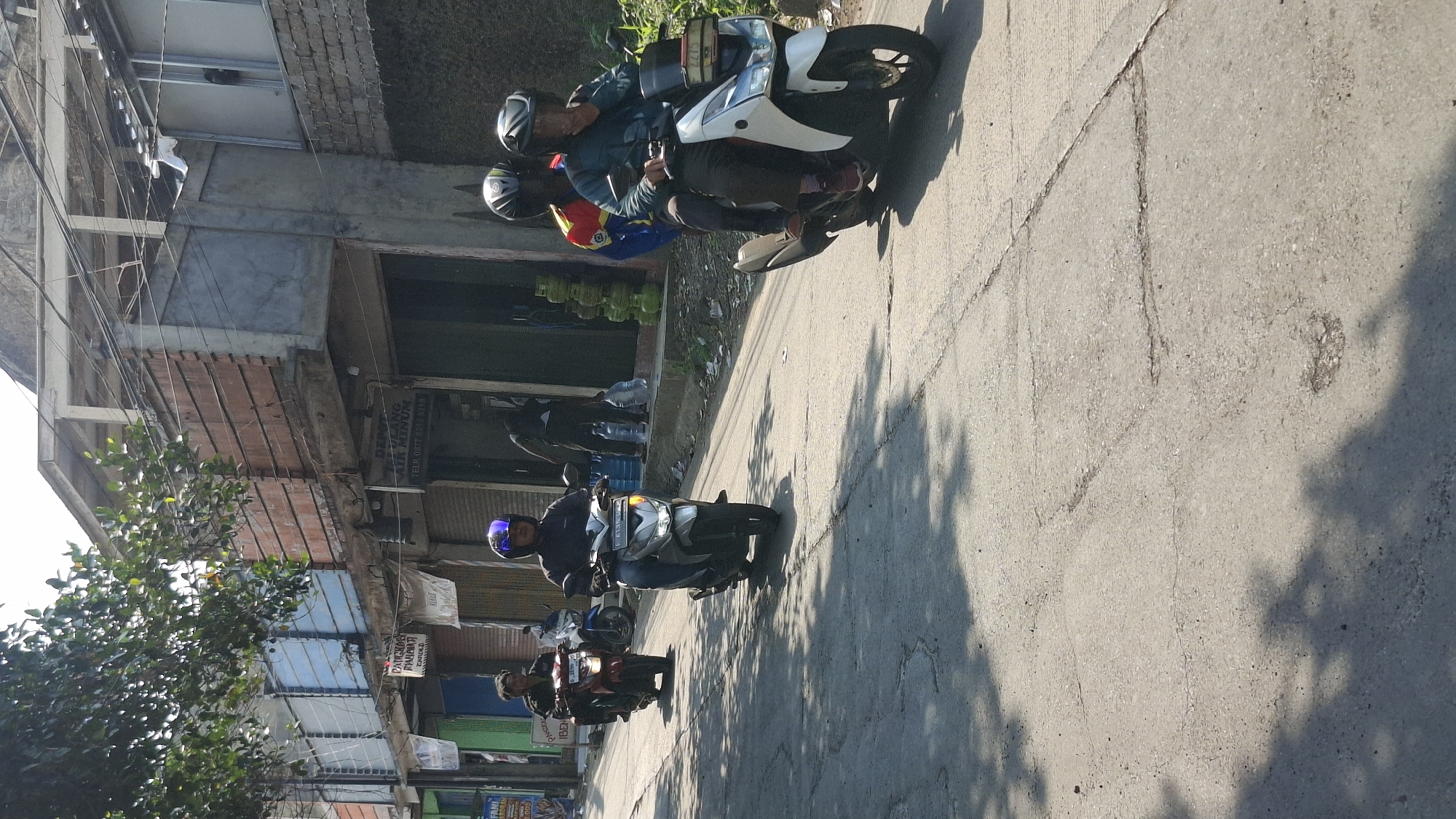 The image depicts a street scene in Cilaku, Indonesia, under bright sunlight casting sharp shadows on the asphalt road. In the foreground, two men on a white motorcycle are riding away from the camera. The driver wears a dark green jacket and dark pants, with a helmet obscuring his face, and has his right hand near his face. The passenger is wearing a yellow and red jacket with a blue collar and a dark helmet. To their left and further down the road, a man on a silver motorcycle, wearing a dark jacket and jeans with a blue and purple helmet, is also riding away from the camera. To his left and even further down, a man on a red motorcycle, wearing a dark jacket and jeans, is also riding away. In the background, a row of buildings lines the right side of the street, featuring a mix of brick and concrete construction. Several shops are visible, one with a sign that reads "DEPOT AIR MINUM" (Drinking Water Depot), displaying large water bottles and gas tanks. Another shop to the left has its door shut and a sign with Indonesian text. Trees with green foliage are visible above the buildings on the left, partially obscuring the sky. The road surface is concrete, showing signs of wear with cracks and some patches. The overall impression is of a typical urban street in Indonesia during the daytime.