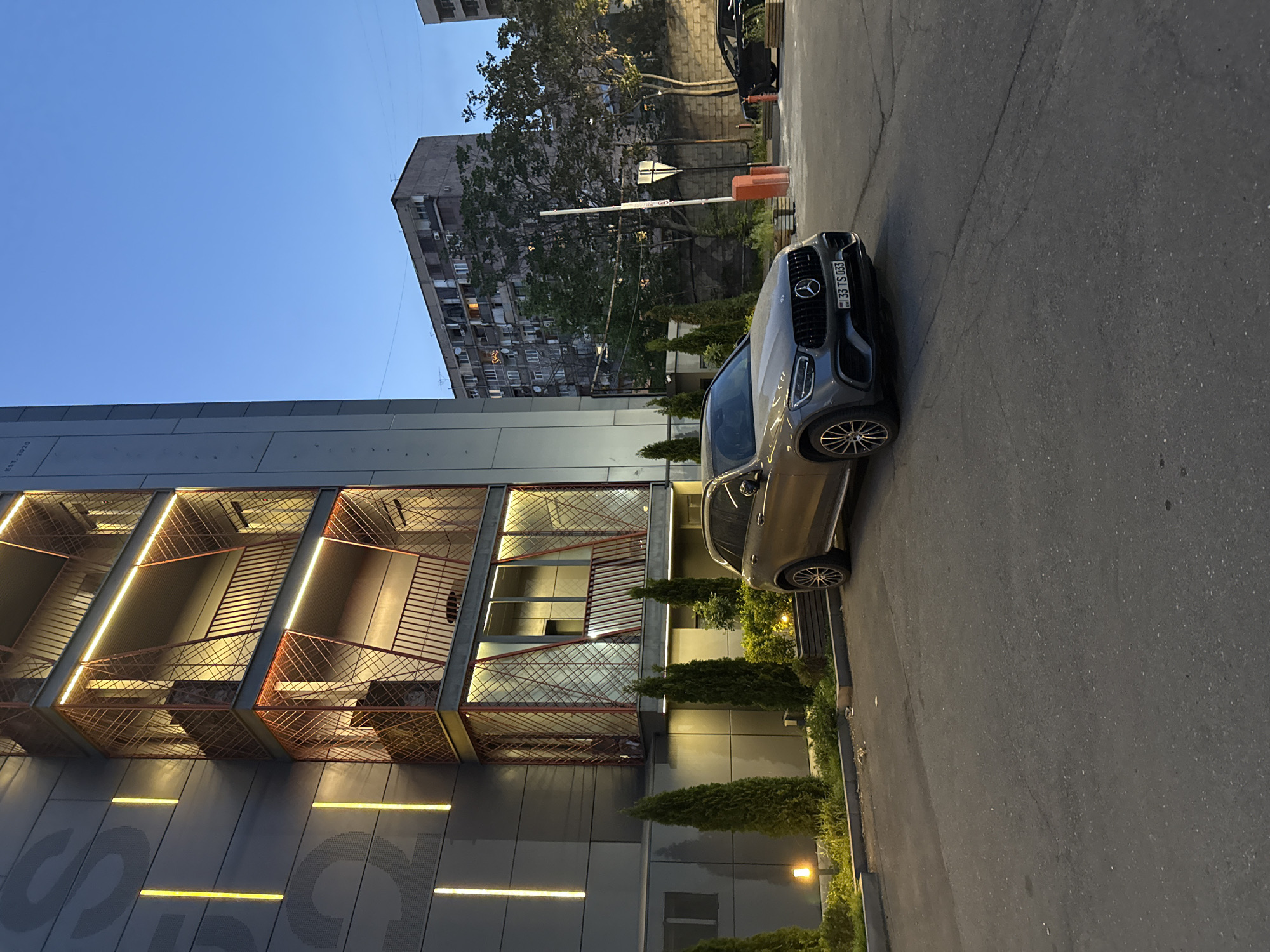 The image shows a gray Mercedes-Benz GLC-Class SUV parked on the side of the road in Yerevan, Armenia. The license plate reads "33 TS 033". Behind the car is a modern building with a unique design featuring exposed staircases with orange railings and reflective glass sections. The building's exterior is gray, with horizontal light bars. In the background, there is an older, Soviet-era apartment building with multiple floors and balconies. The sky is a light blue color, suggesting it is either early morning or late evening. Vegetation in the form of small, cone-shaped trees lines the sidewalk. The road is asphalt and shows signs of wear.
