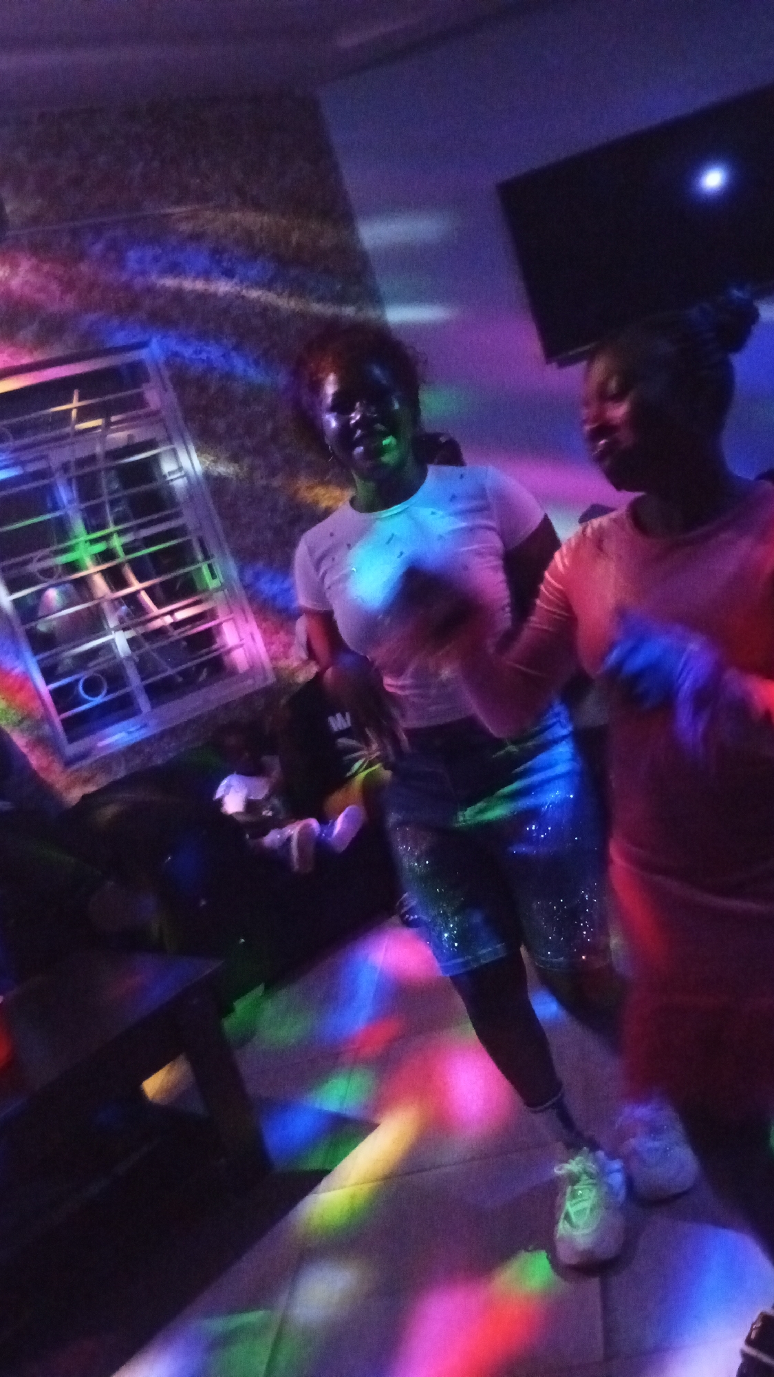 Two young women are dancing and smiling in a dimly lit room illuminated by colorful disco lights. The woman on the left is wearing a white t-shirt and sparkling blue shorts, while the woman on the right is wearing a pink dress. A young child is seated on a couch in the background. The room appears to be a home entertainment area with a television mounted on the wall and a window with blinds.