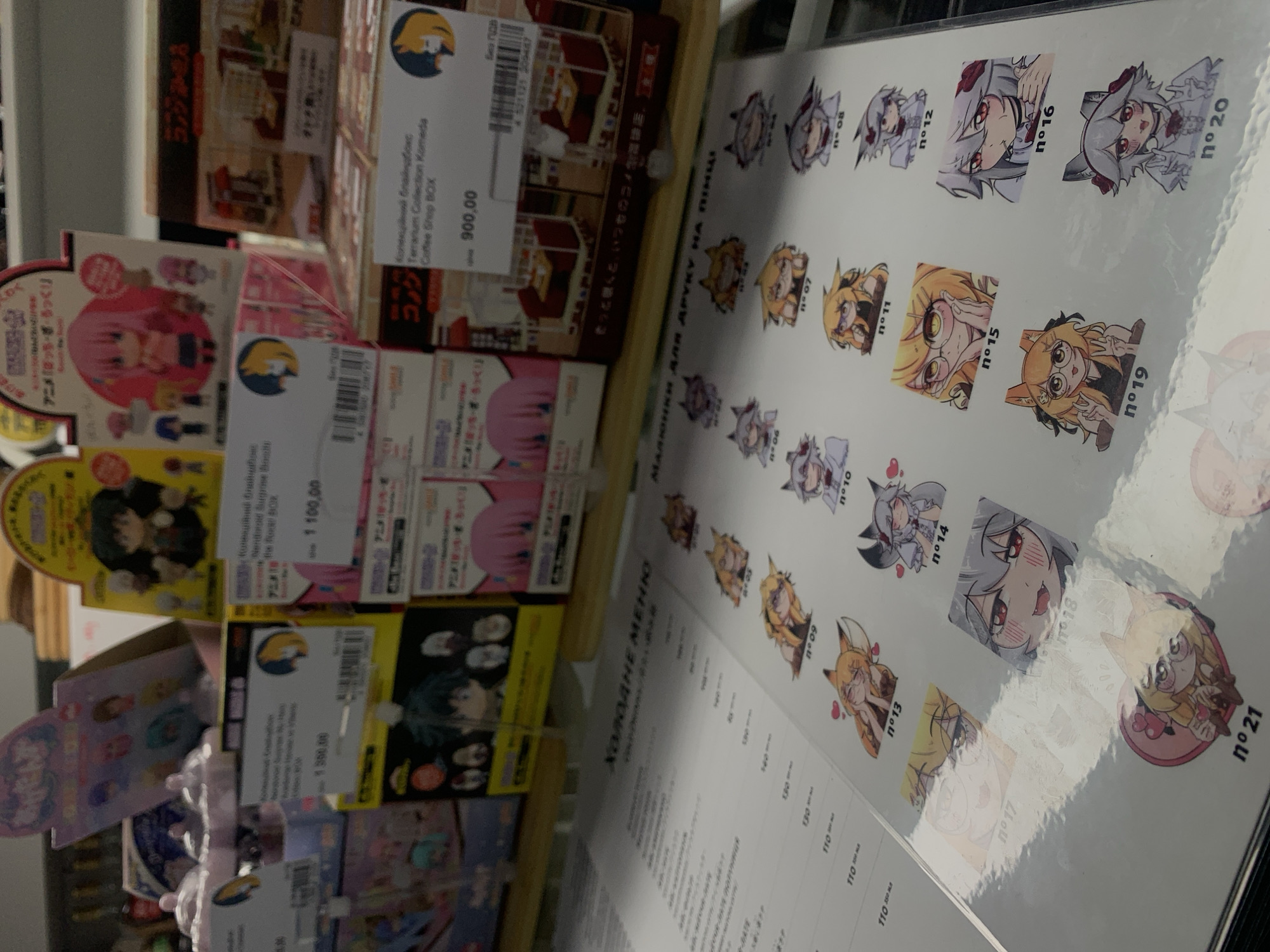 The image shows a display of anime merchandise, possibly in a store. In the background, there are several boxes of collectible figures and toys, some with Japanese text. In the foreground, there is a menu or price list with several small anime character stickers or illustrations, each labeled with a number. The menu is titled in Cyrillic, which translates to "COLD MENU". One of the items on display has a price tag that reads "900,00." The overall scene suggests a retail environment selling anime-related goods. The presence of Cyrillic text indicates a location where this language is used, such as Ukraine, including Kyiv.