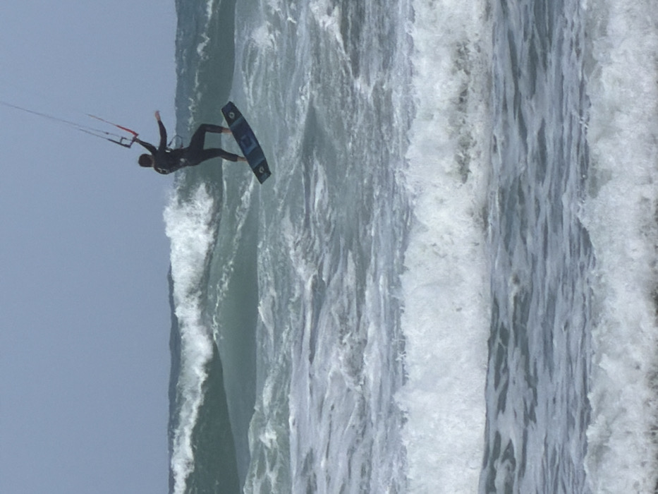 The image displays an individual actively kitesurfing in the Mediterranean Sea off the coast of Tel-Aviv, Israel. The person is suspended in mid-air, wearing a dark full-body wetsuit. A dark blue and black kitesurfing board is affixed to their feet. They grasp a red and black control bar, from which lines extend upwards out of frame, connected to an unseen kite. The water below is rough, characterized by multiple breaking waves, white foam, and choppy surfaces. The sky above is a consistent light grey-blue, indicative of overcast conditions.