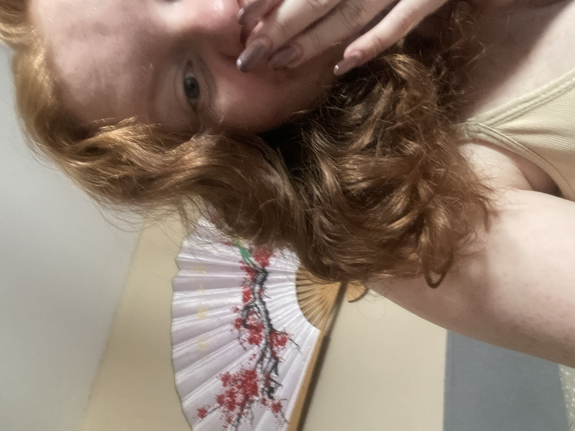A close-up, angled selfie shows a person with reddish-brown, curly hair partially covering their face. Their left eye is visible, and their left hand with long, painted nails is near their mouth. They are wearing a light-colored tank top. A white folding fan with a floral design in red and black is positioned below their hair. The background consists of light-colored walls. The city of Yekaterinburg, Russia, is not visually identifiable in this image.