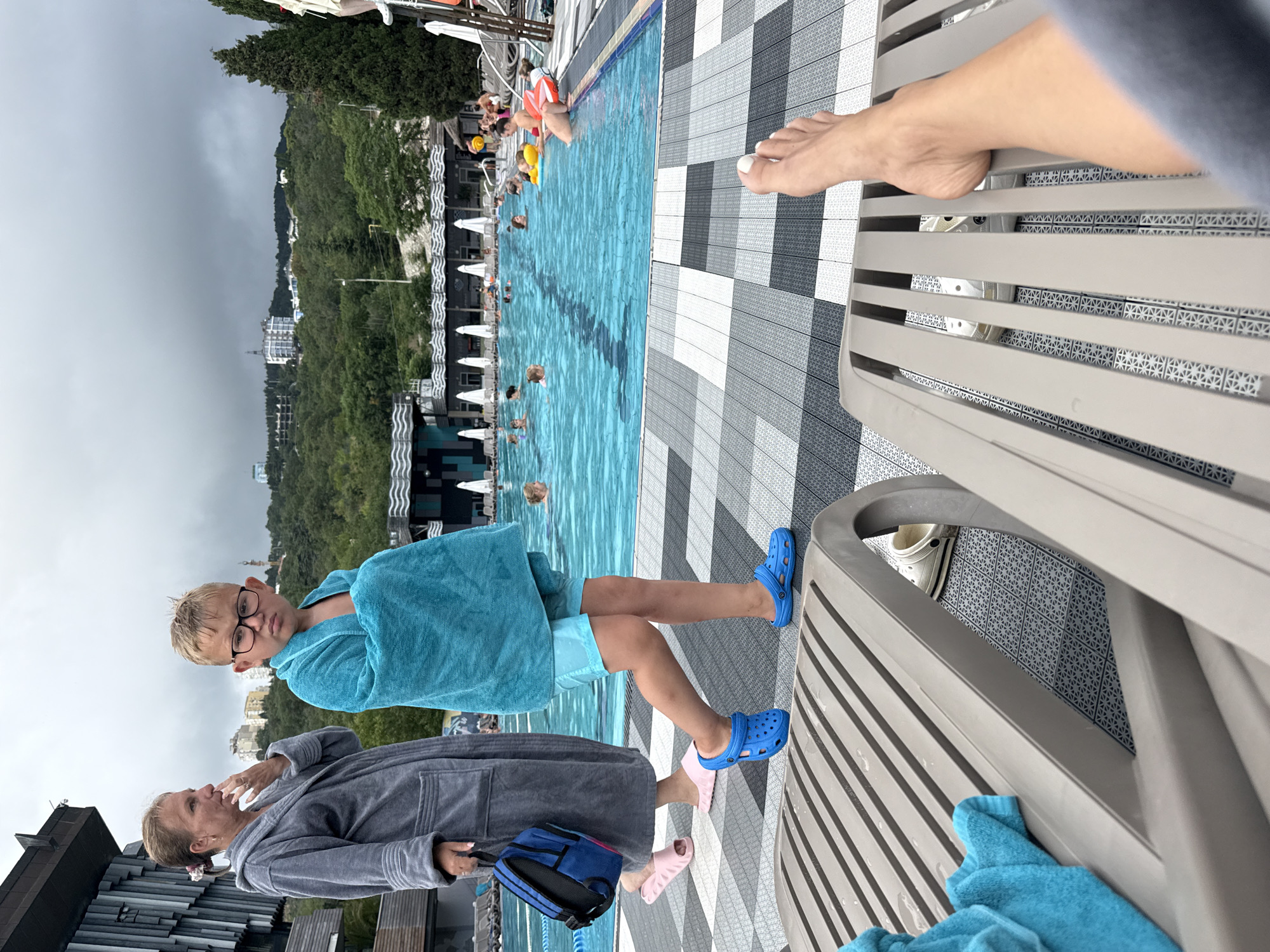 In the foreground, a person's bare feet are visible, resting on a brown lounge chair. To the left of the lounge chair, a blue towel is draped over it. In the mid-ground, a young boy wearing glasses, a turquoise towel wrapped around him, and blue Crocs walks on a patterned tiled walkway. To his left, a woman in a grey bathrobe walks, carrying a blue backpack and wearing pink slides. Behind them is a large outdoor swimming pool filled with people swimming and relaxing. The pool area is surrounded by lounge chairs, umbrellas, and lush green trees, with buildings and hills visible in the background under a cloudy sky. The scene appears to be at a resort or hotel in the city of Yalta, Ukraine.