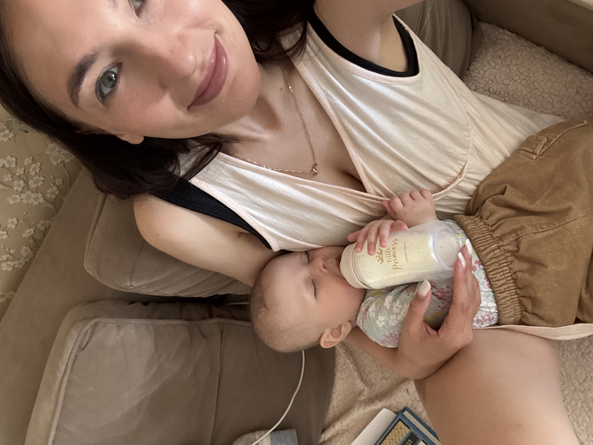 A woman is reclining on a tan-colored sofa with a baby, feeding the baby a bottle labeled "Little Princess." The woman has dark hair, fair skin, and is wearing a light pink v-neck top with a black tank top underneath. She also has a gold necklace. The baby is wearing a light-colored floral-patterned outfit with brown corduroy pants. The bottle is held by a hand with white nail polish. The scene takes place indoors. The city of Sochi, Russia, is not discernible from the provided image.