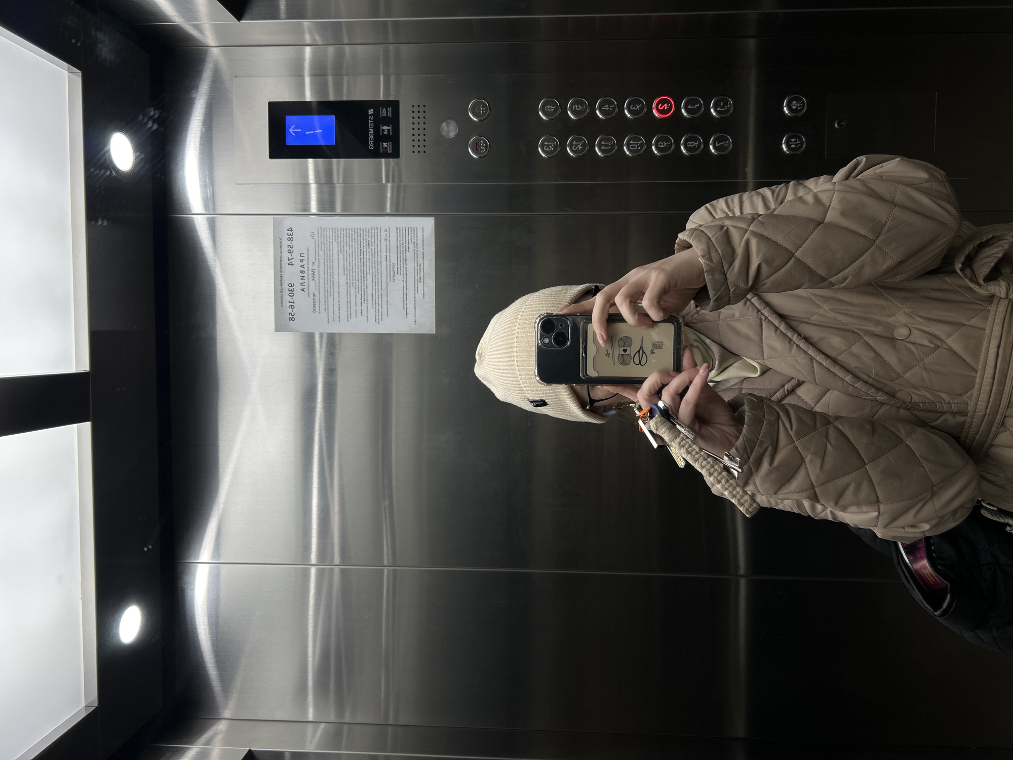 An individual wearing a beige quilted jacket and a knit hat is taking a selfie in a stainless-steel elevator. The elevator panel shows button numbers ranging from 1 to 12, with the number 5 lit up in red. Other panel features include a display screen with an arrow pointing downwards, and an informational sign in Russian. The individual holds a smartphone in front of their face to capture the image. The surrounding environment is that of a modern elevator interior. The location can be determined as Saint Petersburg, Russia due to the Russian language present on the informational sign and elevator panel.