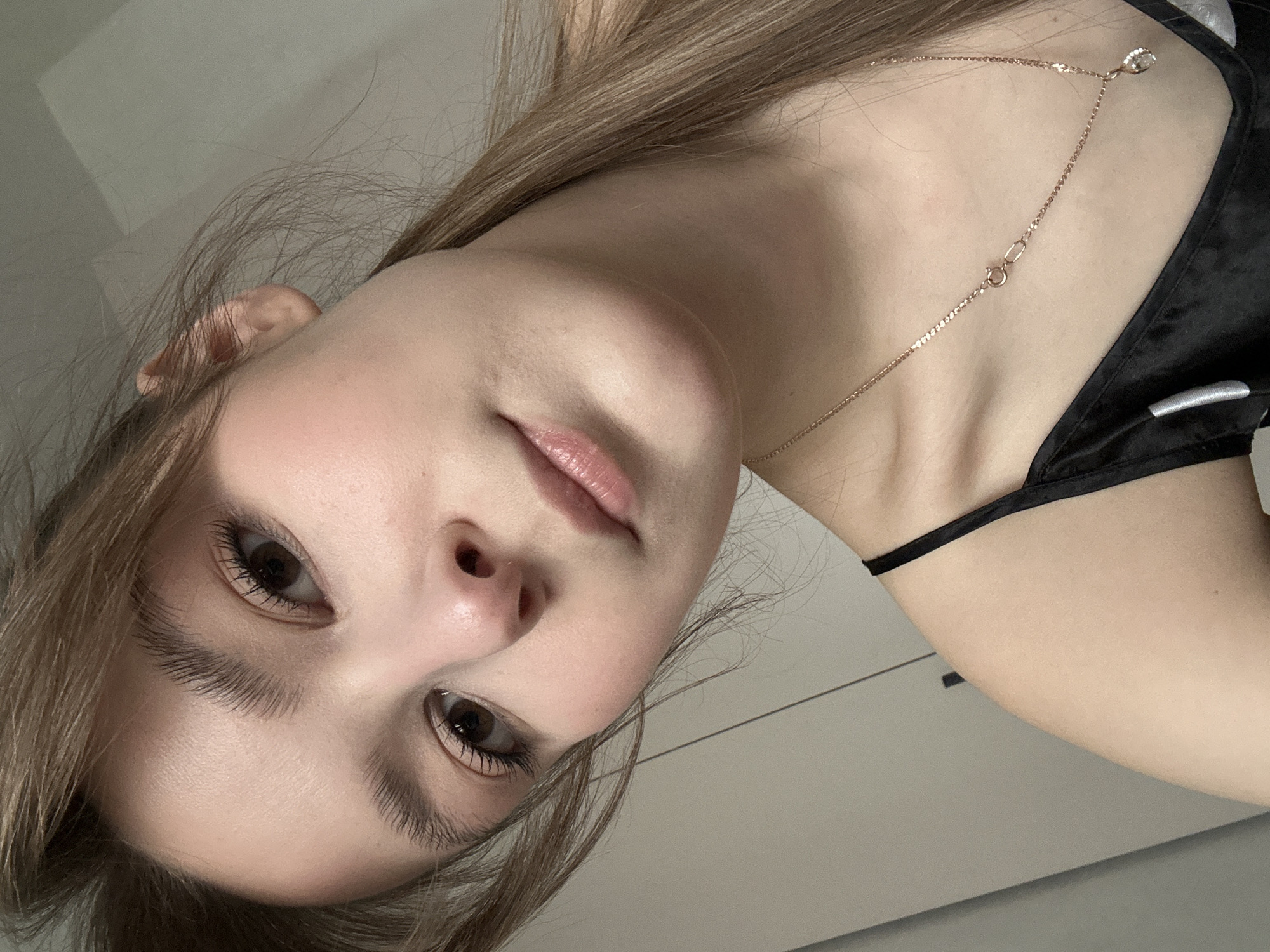 A close-up, rotated portrait captures a young woman with fair skin and long, light brown hair styled loosely around her face. She is wearing a black satin-like top with thin straps. A delicate gold-colored necklace, featuring a small, clear pendant and a visible lobster clasp, adorns her neck. She looks directly at the camera with wide, dark brown eyes, accented by prominent, dark eyelashes. Her lips are full and have a natural pink tint. The background is softly blurred, revealing light, neutral-toned walls or furniture, suggesting an indoor setting. The lighting is soft and even, illuminating her features without harsh shadows. No specific activity is depicted beyond posing for the photograph, and her expression is neutral. There is no visible text. The image was taken in Chaikovsky, Russia.