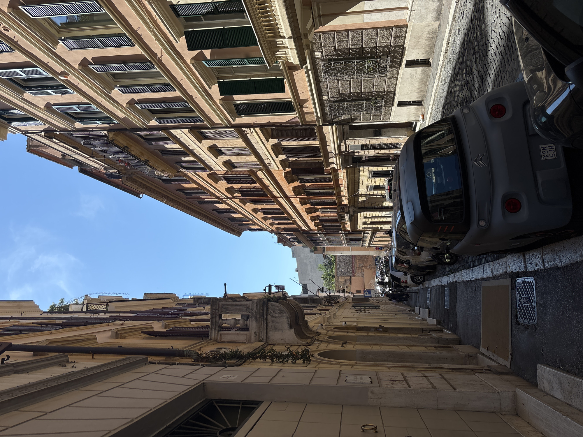 The image shows a narrow street in Rome, Italy, lined with tall buildings on either side. The buildings are old and have many windows with shutters. A variety of parked cars line one side of the street. The pavement is made of cobblestones. The sky is visible at the top of the image and is blue and clear. The overall impression is of a historic, somewhat claustrophobic urban environment. The buildings feature architectural details such as cornices and window frames.