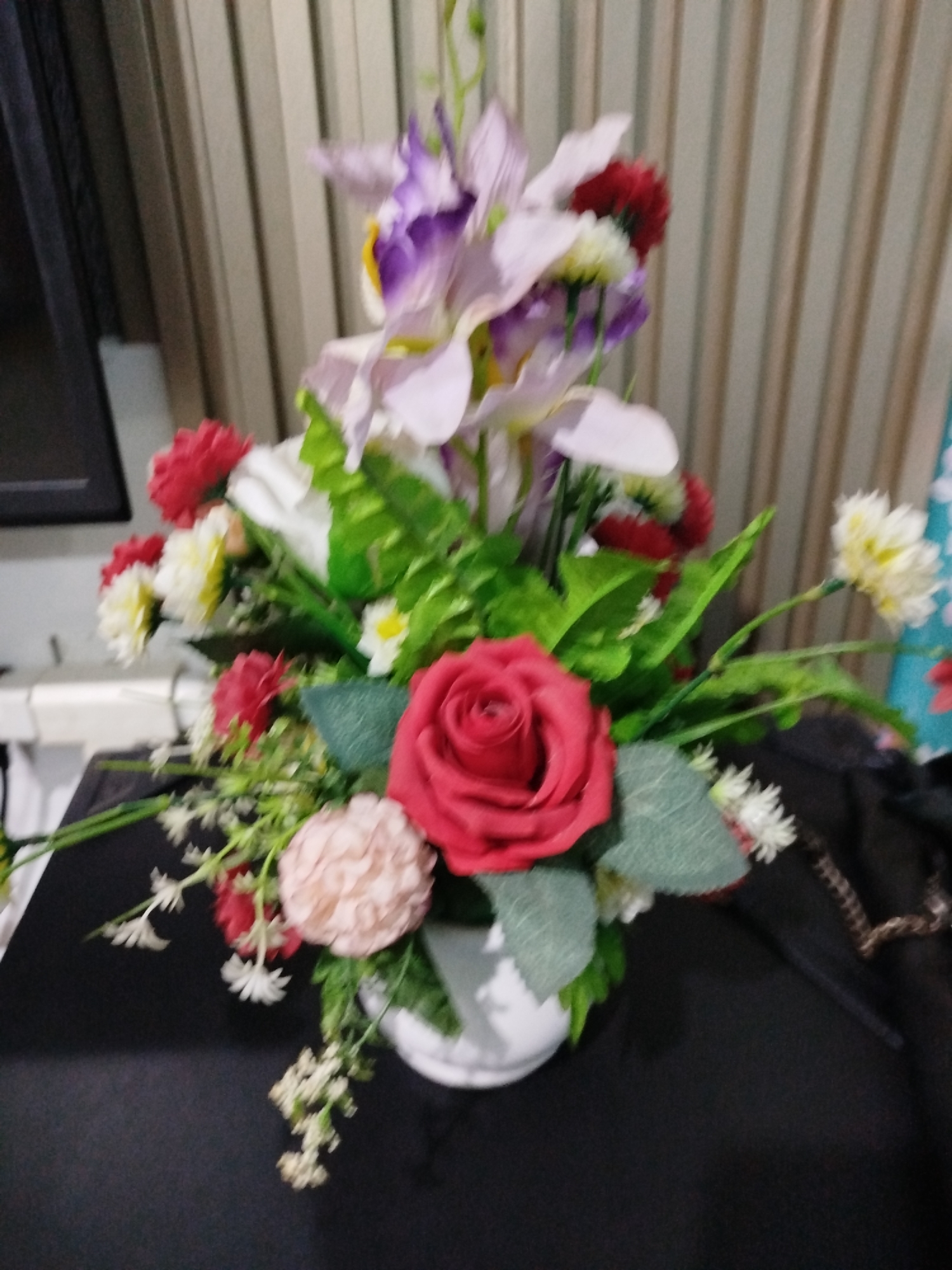 A blurry, full-frame, eye-level shot depicts a floral arrangement in a white vase on a black surface. The arrangement features a prominent red rose, a light pink carnation, and several other flowers in shades of red, white, and purple, interspersed with green leaves. The background consists of vertical, beige blinds. The overall impression is one of indoor decor. The image lacks any indication of Bandungrejo, Indonesia.