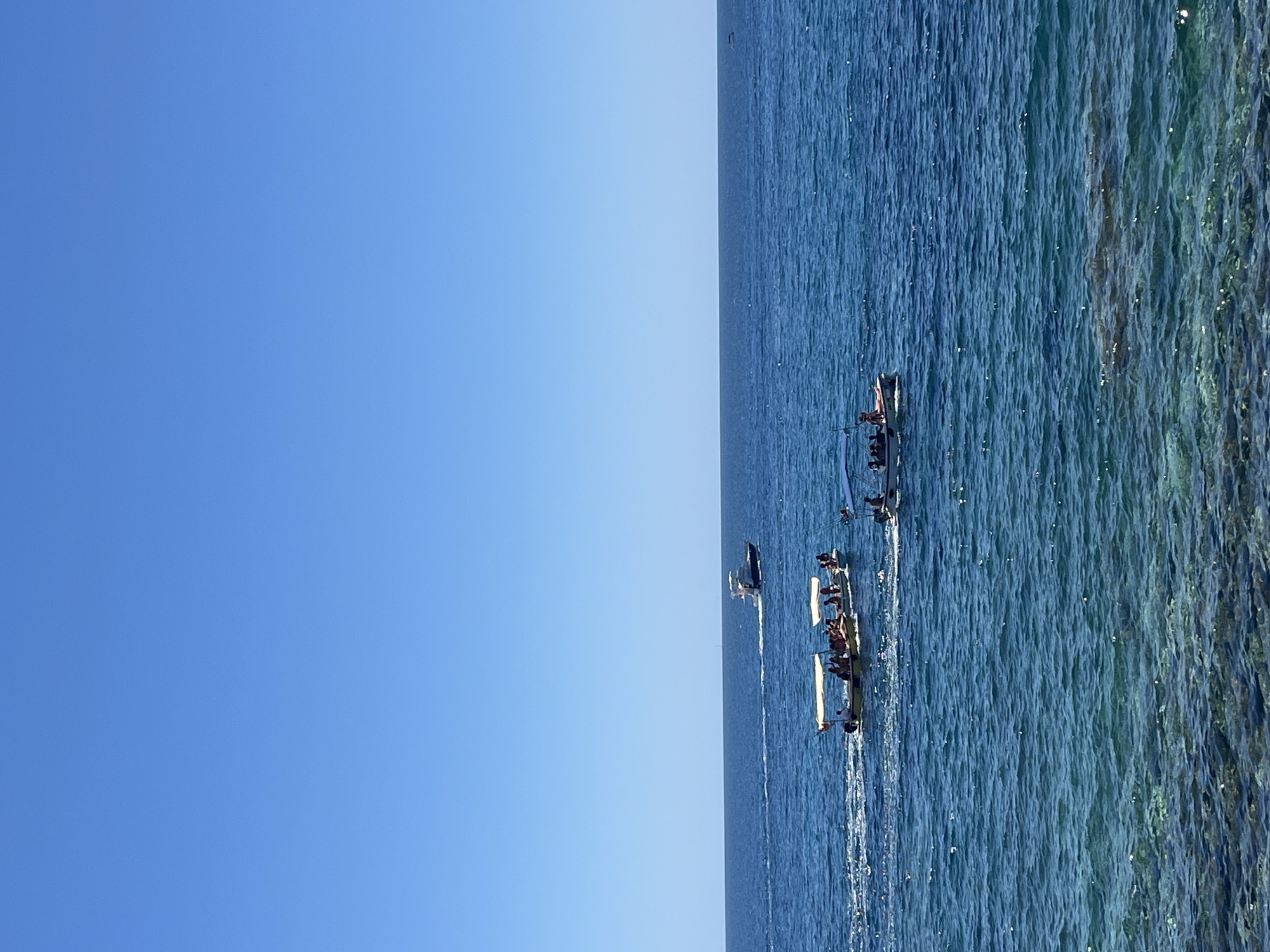 The image shows a sea scene with several narrow boats filled with people moving through the water. The sky is clear and blue, taking up approximately half of the frame on the left. The sea is blue with some areas of lighter, more transparent water closer to the edge. There are three boats visible in the water, moving from left to right. The location is likely near Budva, Montenegro, given the coastal environment. The people in the boats seem to be engaging in a recreational activity, possibly a tour or race.