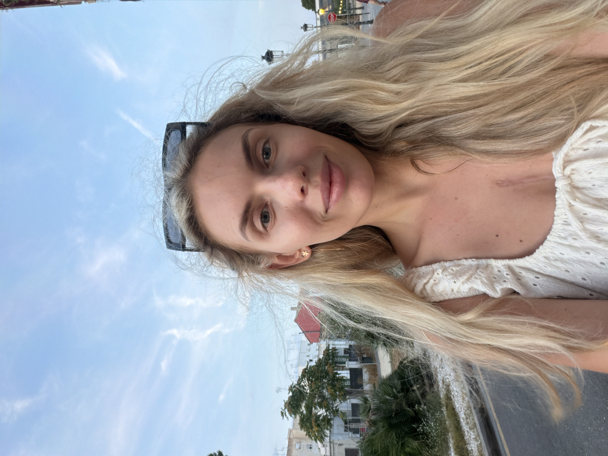 A light-skinned woman with long blonde hair and sunglasses perched on her head smiles at the camera. She is wearing a white eyelet top with ruffled sleeves and a tie in the front. A scar is visible on her chest. The background shows a street with trees and buildings, and a stop sign is visible in the distance. The location is the Benimàmet-beniferri district of Valencia, Spain.