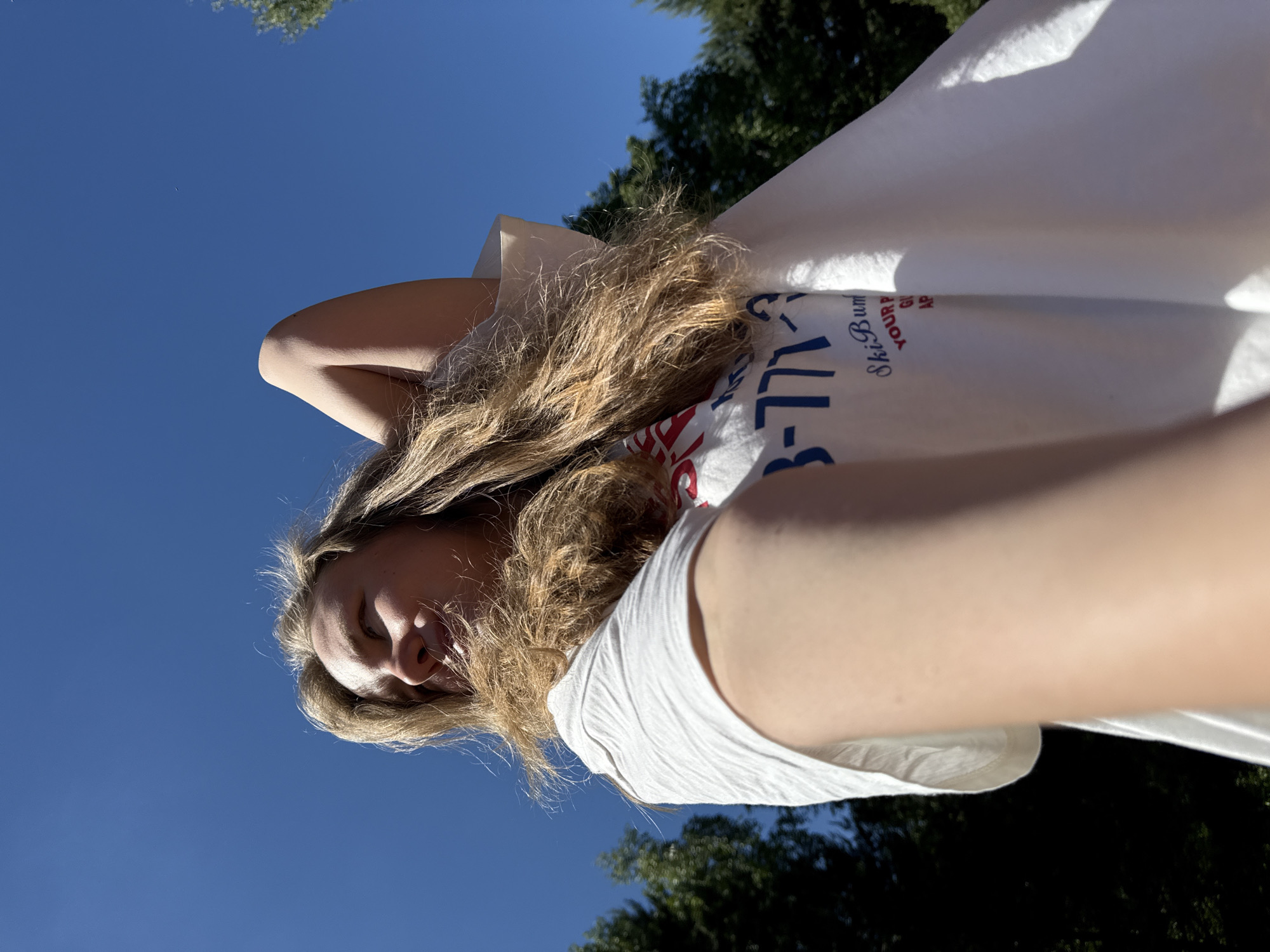 A low-angle shot captures a person with long, wavy blonde hair looking upwards and to their left. Their right arm is bent, with the hand positioned behind their head. They are wearing a white short-sleeved t-shirt with partial text visible, including "3-771-3" (likely a phone number) and "Ski Bum," followed by fragments such as "YOUR P," "GI," and "AP." An outstretched arm, possibly the subject's left arm holding the camera, partially obscures the lower-right foreground. The background is dominated by a clear, bright blue sky, with dark green foliage from trees visible along the left and right edges of the frame, indicating an outdoor setting under direct sunlight. The image provides no visual indicators to confirm the specific location within the city of Копнино, Russia.