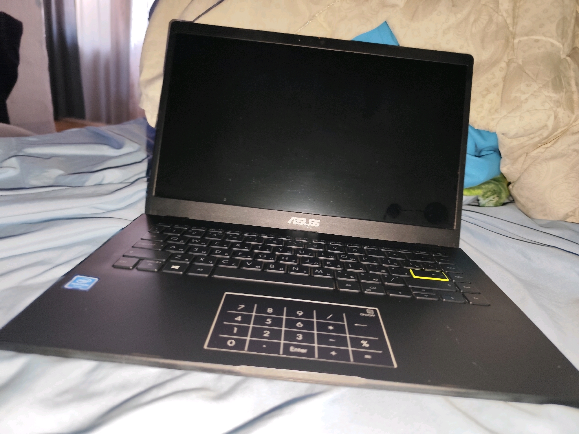A black ASUS laptop is open and angled towards the viewer, resting on a bed with blue and white patterned sheets. The laptop screen is dark, reflecting some light. The keyboard is visible, with a yellow key prominently displayed. Below the keyboard is a touchpad with a numbered keypad overlay. A blue and white sticker, likely a brand logo, is on the left side of the laptop's base. The background shows draped bedding and curtains, suggesting an indoor setting. The image does not contain any information about the location of Balqaş, Kazakhstan.