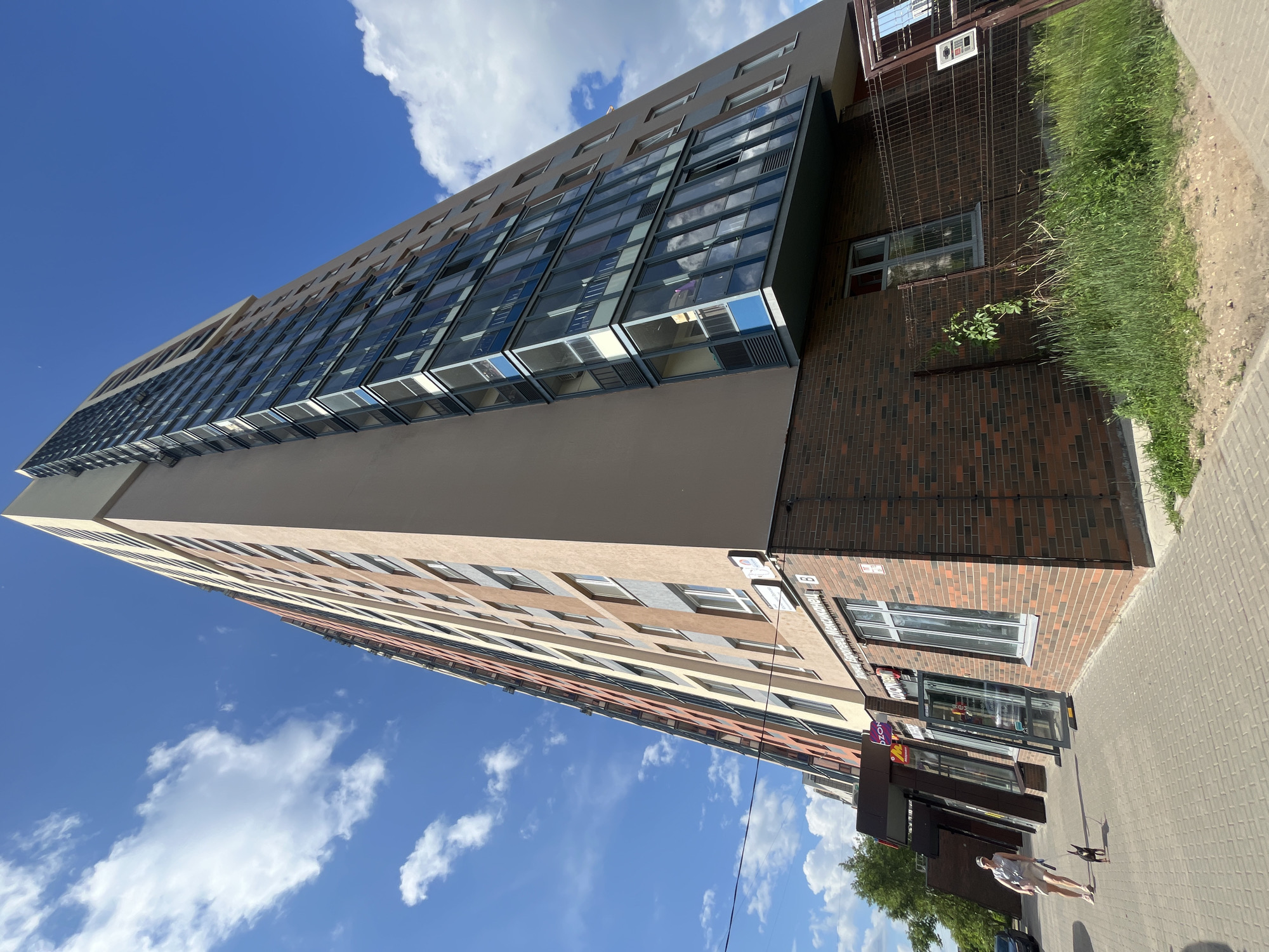 A low-angle, full-shot photograph captures a tall, modern apartment building on a sunny day in Perm, Russia. The building's facade is a mix of light tan, grey, and vertical bands of orange and yellow. The lower portion of the building is clad in dark red brick, featuring large glass-fronted shops on the ground floor. To the left, a woman wearing a light-colored shirt and shorts walks her small black dog along the paved sidewalk. The sky is a vibrant blue with scattered white clouds. A metal fence encloses an area with overgrown green grass and weeds in front of the building's brick base. The image appears to be taken on a street in a residential or mixed-use urban area.