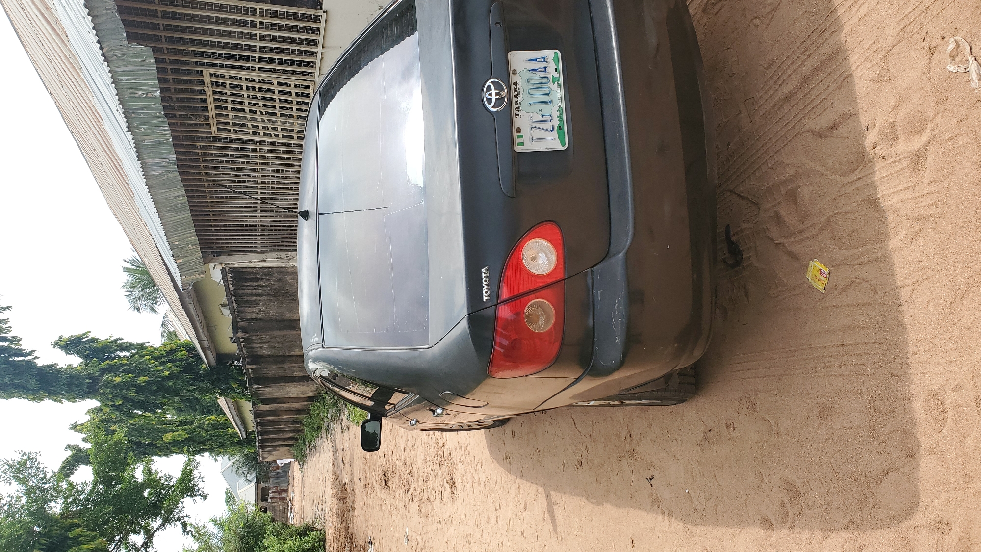 In Jalingo, Nigeria, a dark-colored Toyota sedan is parked on a dirt road, partially obscuring a residential building with a corrugated metal roof and barred windows. The car's rear is visible, featuring a Toyota logo and a license plate that reads "TZG 100AA" and "TARABA." Tall, slender trees line the left side of the road, casting shadows on the sandy ground. The environment appears to be an outdoor residential area, with evidence of vehicular movement on the dirt surface.