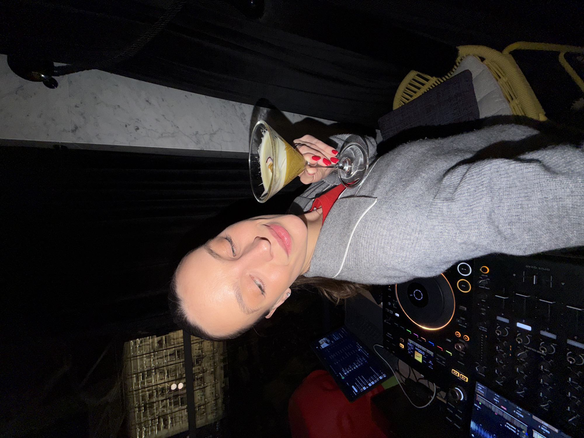 A woman with closed eyes holds a martini glass filled with a yellow liquid in her right hand, while resting on a black DJ console with a laptop nearby. The woman wears a grey sweater with a red collar. Her fingernails are painted red. A building with lit windows is visible in the background, and part of a yellow wicker chair is also visible. The location is the Top Hill club in Budva, Montenegro.