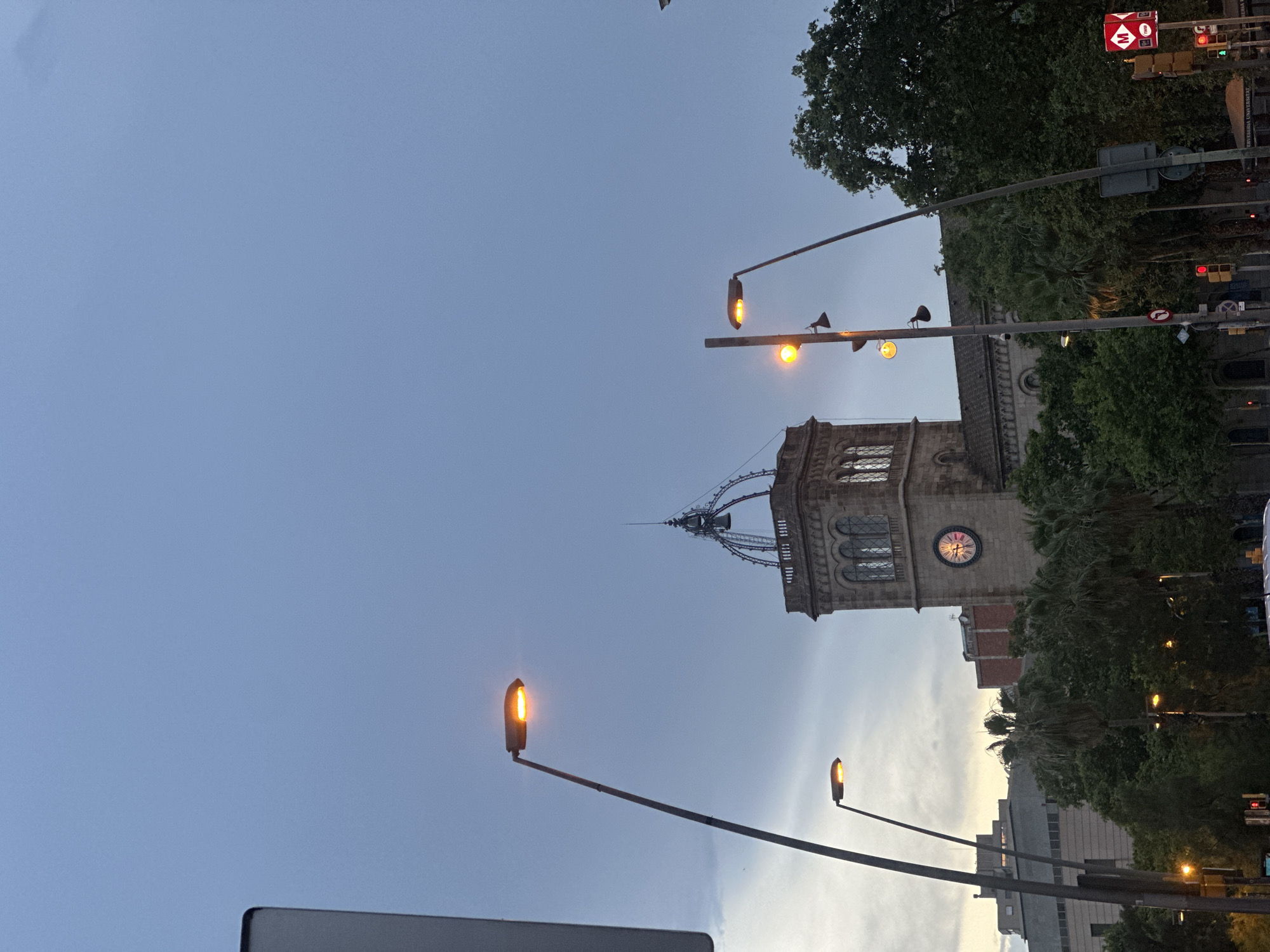 The image shows a view of the University of Barcelona from the street, likely during twilight. A prominent clock tower with a dome is visible in the center, flanked by trees and modern buildings. Several streetlights are illuminated, casting an orange glow. Traffic lights and signage, including a red metro sign, are present at the street level. The sky is a muted blue, suggesting the end of the day.