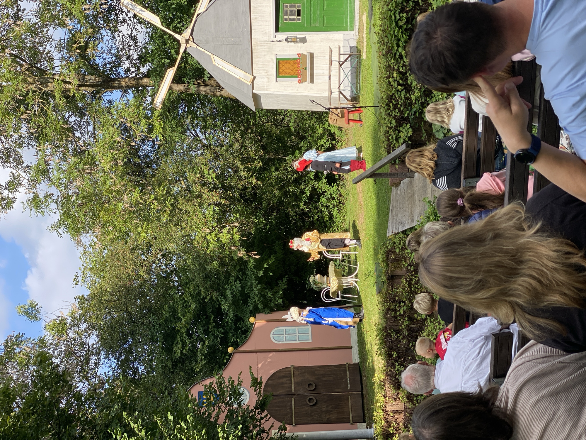 The image depicts an outdoor performance on a grassy stage in Flammersfeld, Germany, likely at a park or festival. In the background, a pink building with large wooden doors and a blue emblem on its facade stands to the left, and a white windmill structure with a green door is to the right. Four individuals are on the stage, dressed in costumes. Two are in the foreground of the stage: one in a blue outfit with white rabbit ears and a staff, and another in a golden robe with a crown and a beard. Further back, two figures in blue and red costumes are also present. A small table and chairs are set up on the stage. In the foreground, an audience is seated on wooden benches, viewed from behind. The environment is characterized by lush green trees and a partly cloudy blue sky.