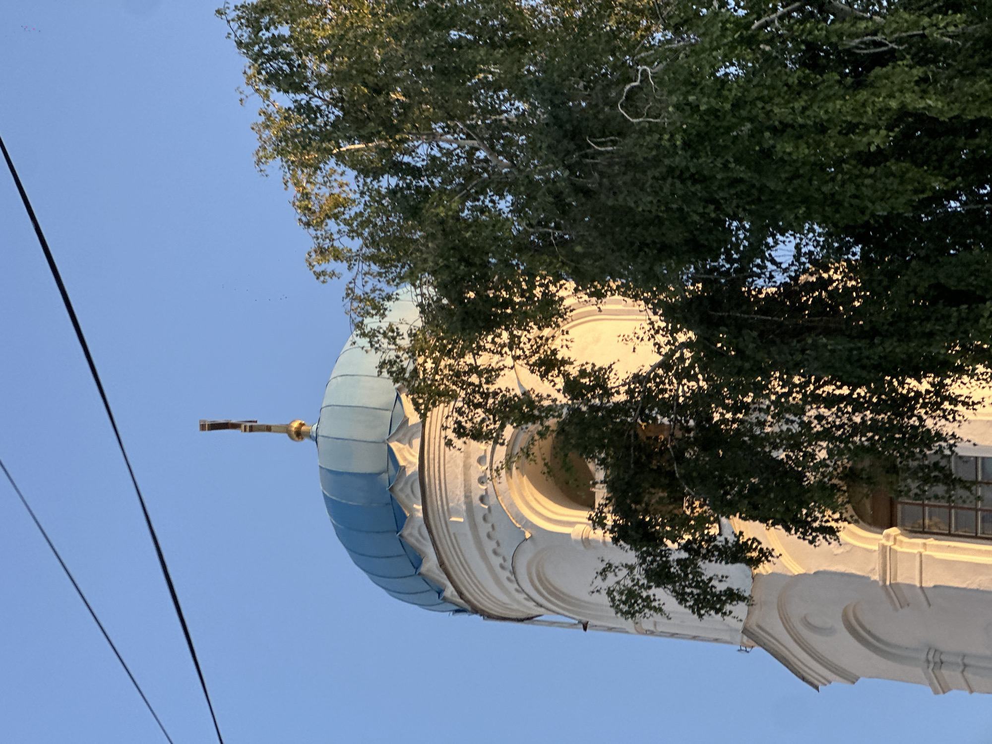 A medium-shot, low-angle outdoor photograph captures the upper section of a white bell tower topped with a blue dome and a gold cross. The bell tower features arched openings and decorative moldings. A lush green tree with dense foliage partially obscures the bell tower, with sunlight illuminating the side of the tower and parts of the tree. Two dark power lines are visible diagonally across the top of the clear blue sky. The scene is likely a religious building in Biysk, Russia, given the architectural style and the presence of a cross. No people are visible in the image.