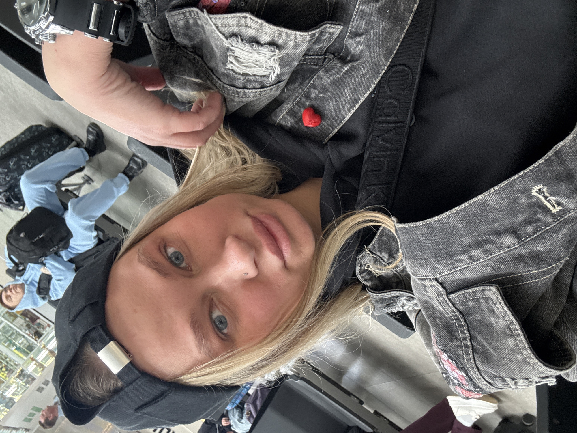 A selfie of a person with fair skin, blonde hair, and blue eyes, wearing a black hooded sweatshirt under a gray denim jacket with distressed details and a small red heart pin. They have a piercing on their nose. A black Calvin Klein-branded strap is visible across their chest. They are wearing a black digital watch. In the background, there is a person sitting with light blue clothes and luggage. The floor appears to be light gray tile. The location of Boldino, Russia is not apparent within the visible confines of this photograph.