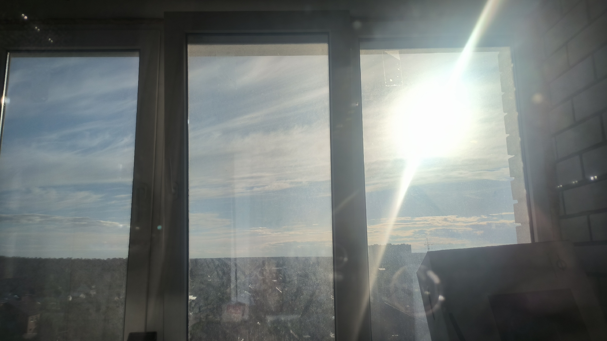 The image shows a view from a window in Kovrov, Russia, looking out at a cityscape under a bright sun. The window is divided into three panes, with the left and middle panes showing a sky with wispy clouds and a distant treeline. The right pane is dominated by the sun, which is shining intensely and creating lens flare and glare across the glass. The exterior of the building to the right of the window is visible, with white brickwork. The bottom part of the image shows more of the city, with buildings and what appears to be a forest or park area. The overall impression is a sunny day with a clear view of the horizon.