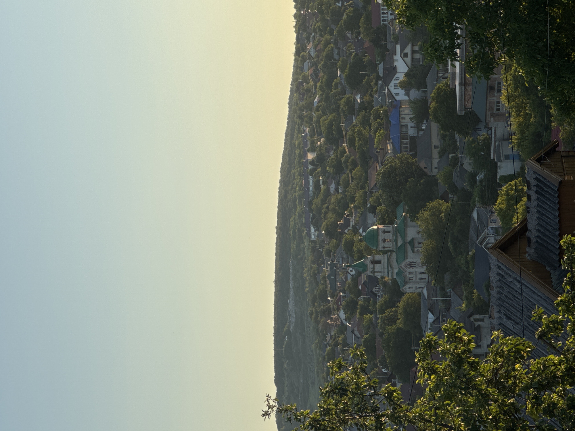 The image captures a panoramic view of a residential area in Strășeni, Moldova, nestled amidst rolling hills. In the mid-ground, a prominent white church with green domes stands out against the dense foliage of trees that surround and dot the urban landscape. The scene is a compilation of houses with red, grey, and blue roofs, interspersed with lush greenery, suggesting a rural or semi-rural setting within the city. The foreground features the tops of wooden structures with corrugated roofing, adding a rustic element to the composition. The sky above is a gradient of soft blue and pale yellow, indicative of either early morning or late afternoon light. No people are visible in the image. The overall impression is one of tranquil, green residential sprawl.