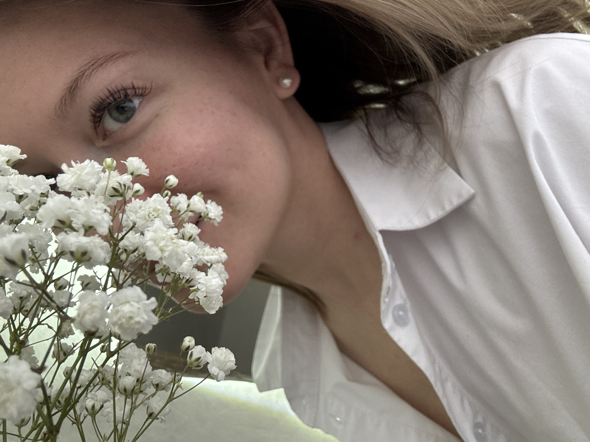 A close-up, low-angle shot features a young woman with light skin and freckles partially obscured by a bouquet of white baby's breath flowers. Her left eye, with prominent eyelashes, is visible. She wears a white, collared shirt, partially unbuttoned at the neckline. A small, pearl earring is visible in her left ear. The background is out of focus, with a hint of hair and possibly window bars. No specific location within Chaikovsky, Russia, can be determined from this image.