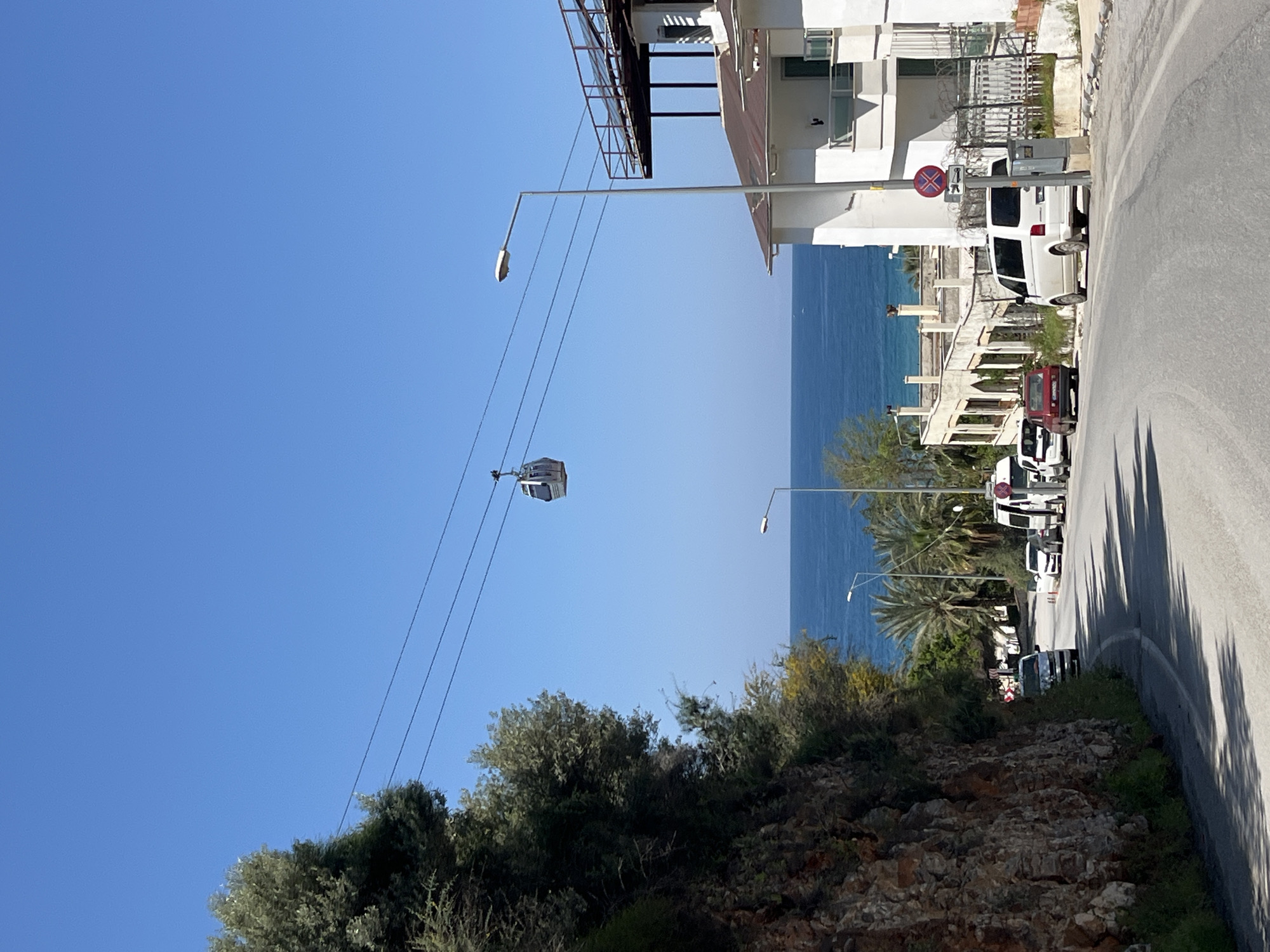 The image depicts a coastal street scene in Alanya, Turkey. The view looks down a road alongside the sea. On the right of the road, multiple parked vehicles are visible, including white vans and a red car. A building stands near the road, with a balcony overlooking the sea. A "no pedestrian crossing" traffic sign is attached to the building. On the left, a cliff covered in vegetation slopes down towards the beach, obscuring the view. Overhead, a cable car system runs across the scene, with one car visible in the air. The sky is clear and blue. Streetlights are also visible alongside the road.