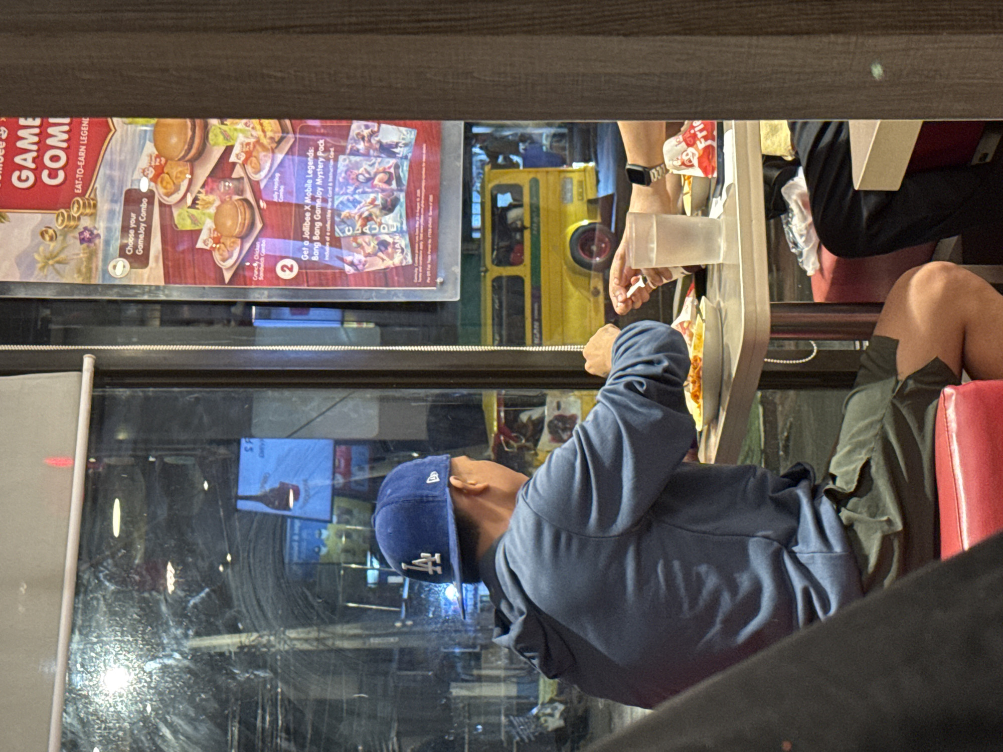 The image, originally captured in a portrait orientation but depicting a horizontal scene, shows two individuals eating inside a restaurant next to a large window. In the foreground left, a person wearing a blue hoodie and a blue baseball cap with a white "LA" logo is seated, looking down at food on a light-colored table. To the right, another person's hand and lower arm, wearing an Apple Watch, are visible, holding a plastic cup. Part of this person's leg, in dark shorts, rests on a red upholstered seat.
Outside the window, a blurred street scene is visible, including a distinctive yellow vehicle, likely a jeepney, suggesting a Philippine setting. The time of day appears to be evening or night, indicated by the dark exterior and prominent indoor reflections on the window.
Above the window, a large advertisement is displayed. Visible text includes "GAME COMBO," "EAT-TO-EARN LEGEND," "Choose your GameJoy Combo," "Crunchy Chicken Sandwich Combo," "Jolly Hotdog Combo," and "Get a Jollibee X Mobile Legends: Bang Bang GameJoy Mystery Pack," which "Includes a collectible Rare Card & Instruction Card." The ad features images of burgers, hotdogs, and fries. This advertisement, combined with the location context of Davao City, Philippines, strongly indicates the establishment is Jollibee.