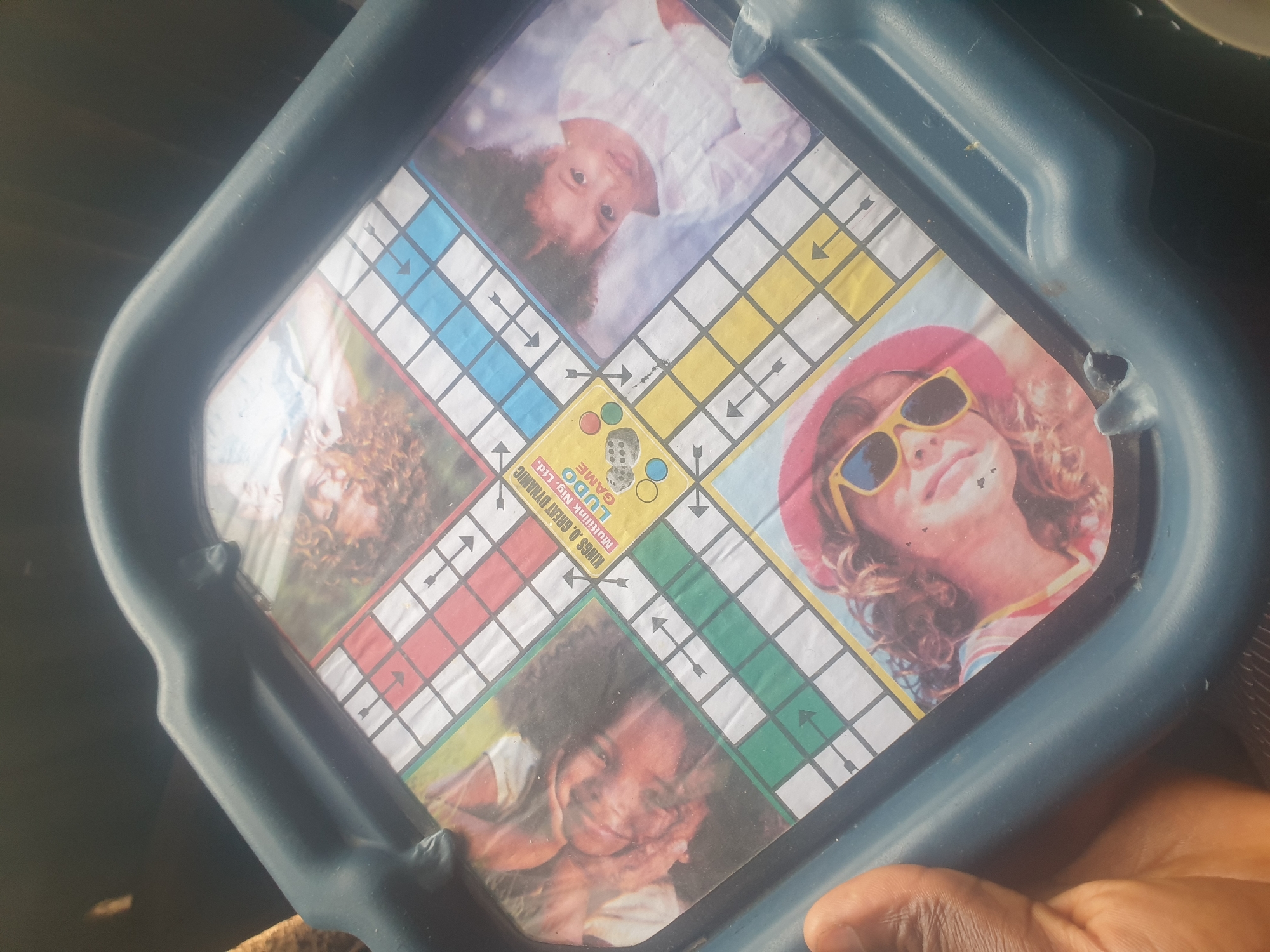 A Ludo game board is shown, centered within a blue plastic tray. The game board features a colorful design with pathways marked by squares in red, blue, green, and yellow. In the center of the board, there is a logo that reads "KINGS O. GREAT DYNAMIC LUDO GAME." Four quadrants of the board are adorned with portraits of children and adults. The top left quadrant shows a person's face with curly hair. The top right quadrant features a young child with curly hair, wearing a white outfit. The bottom left quadrant displays a person with a hat and sunglasses. The bottom right quadrant shows a young child, also with curly hair. The game board appears to be placed outdoors, with ambient light suggesting a daytime scene. The image is taken from a high angle, with a portion of a person's hand visible in the lower left corner. The overall setting implies a casual or recreational context. The image context provided by the user states the location is in the city of Jos, Nigeria.
