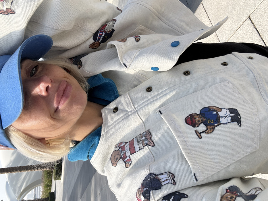 A woman with short blonde hair and a blue cap poses in front of two light-colored denim jackets. Each jacket has multiple patches of teddy bears dressed in various outfits. She wears a blue hoodie under the jackets. The background shows a blurred outdoor scene with light-colored pavement and architectural elements.