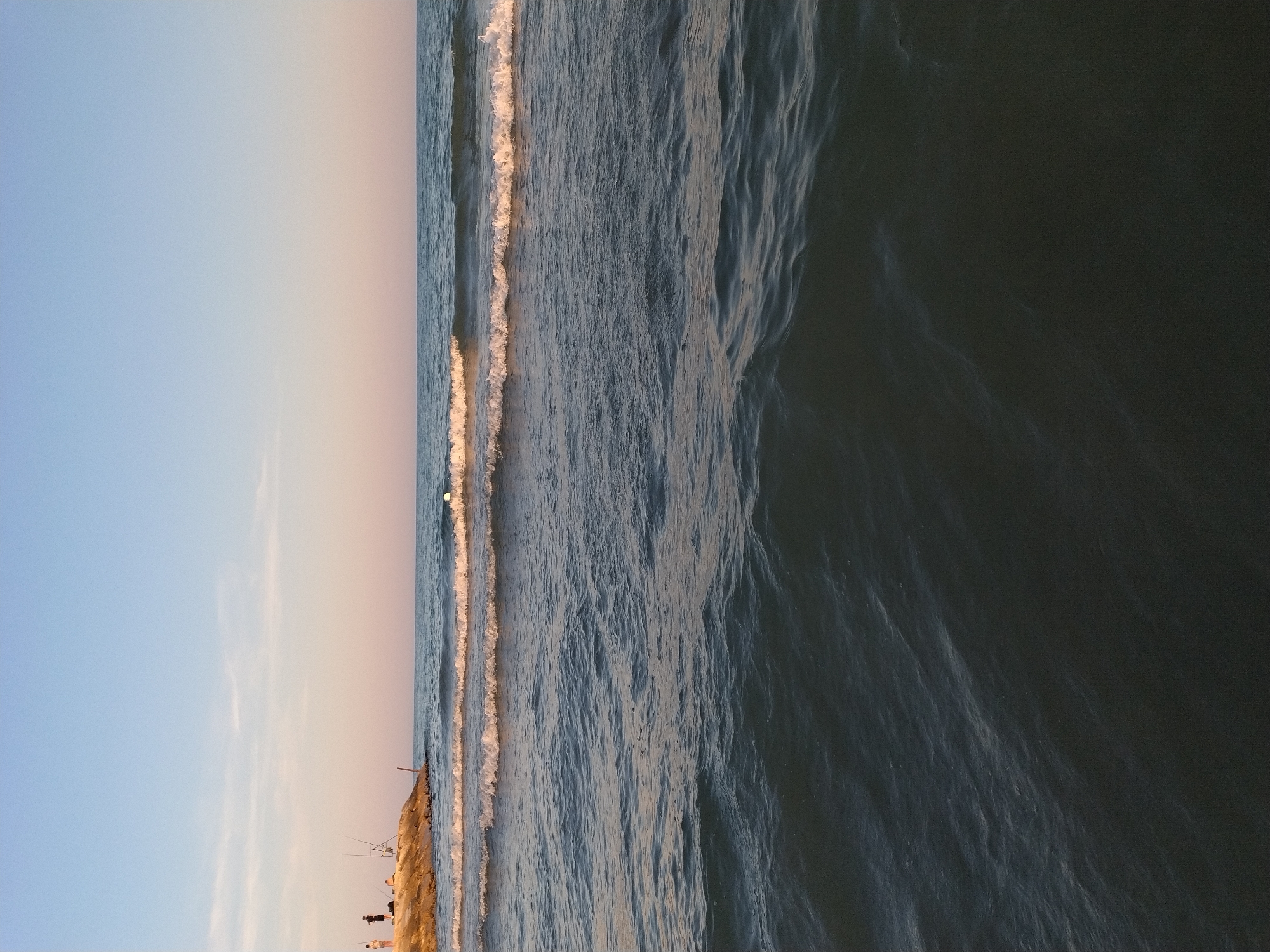 The image depicts a serene coastal scene in Caorle, Italy, at what appears to be dusk. In the background, a rocky embankment or breakwater juts into the sea, upon which three individuals are visible. Two of them appear to be standing, while a third person is seated. Fishing rods are positioned on the embankment, suggesting recreational fishing is taking place. The sea is calm with gentle waves rolling towards the shore, creating white foam along their crests. The sky exhibits a gradient of colors, transitioning from a clear blue at the top to a soft pinkish hue near the horizon, characteristic of sunset or sunrise. The foreground is dominated by the dark, rippling surface of the water, reflecting the subdued light of the sky.